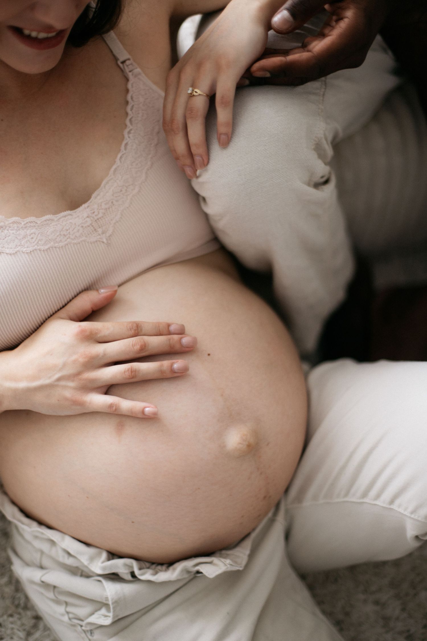 Pregnant woman in beige lace bralette cradling her bare baby bump while partner places hand on her shoulder.
