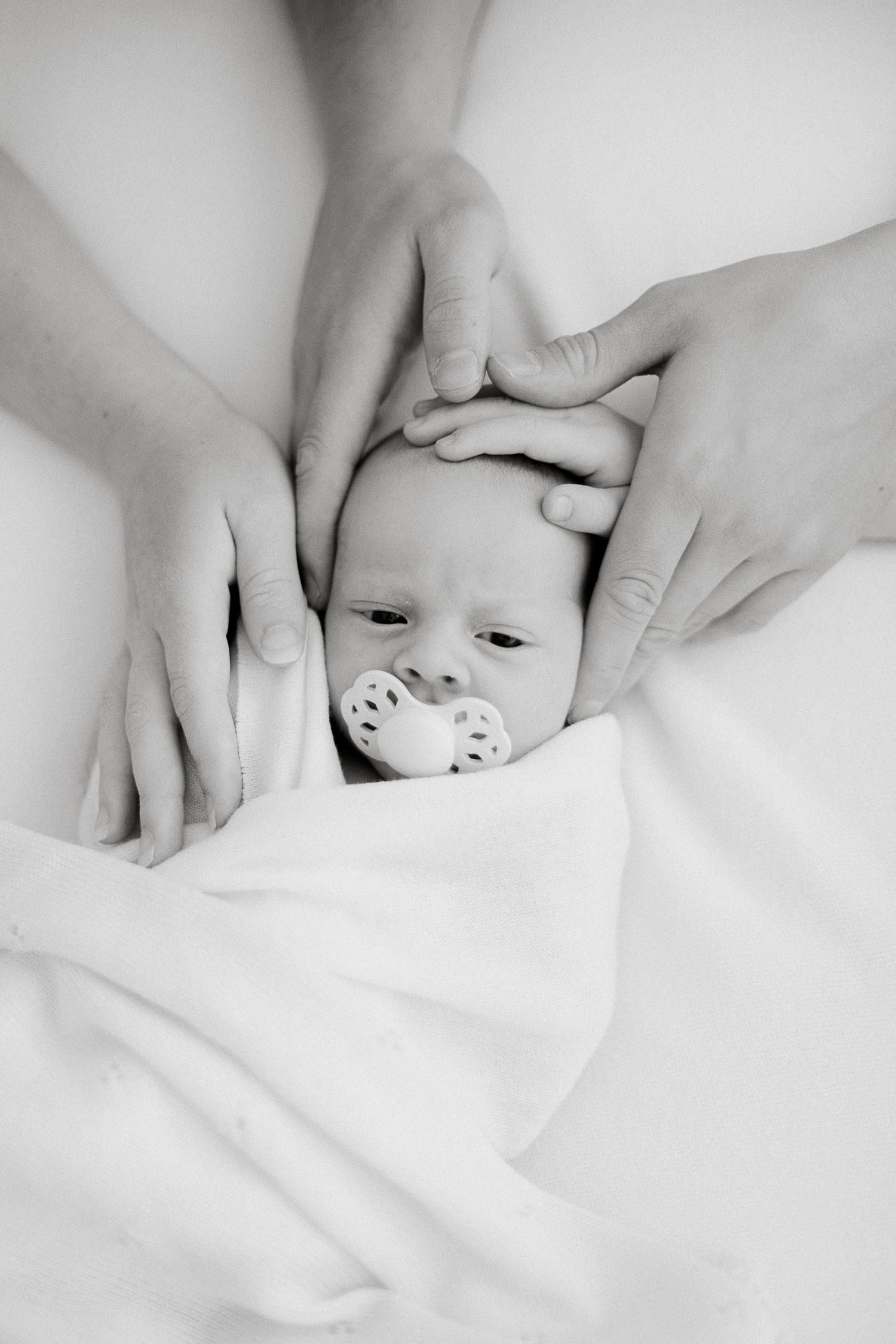 Black and white photo of a newborn baby with a pacifier, cradled gently by two pairs of adult hands on a white blanket.