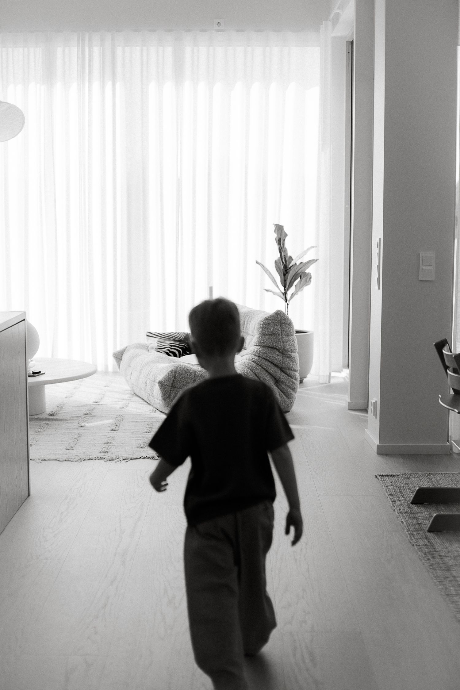 Black and white photo of a young boy walking through a modern living room toward bright curtained windows.