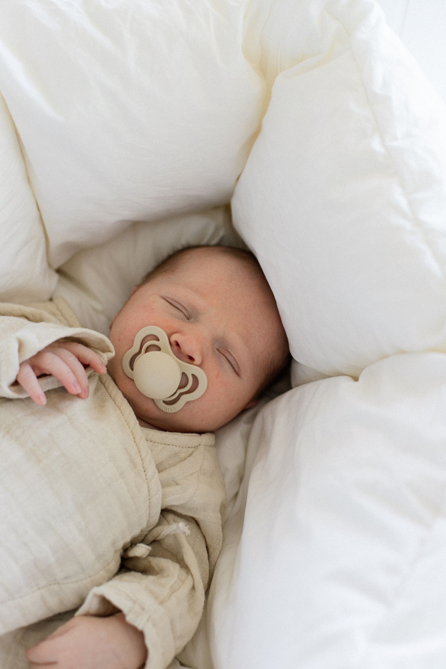 Peaceful newborn baby sleeping with a pacifier, wrapped in a beige outfit, nestled among soft white pillows.