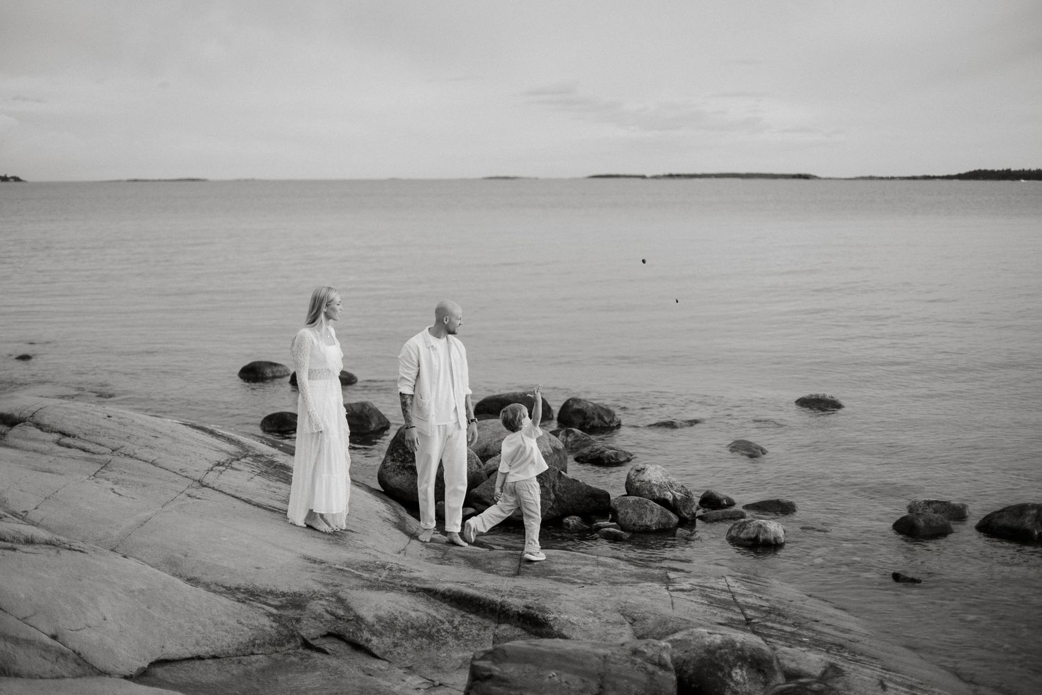 Family of three standing on rocky shoreline gazing at calm sea in black and white photo.