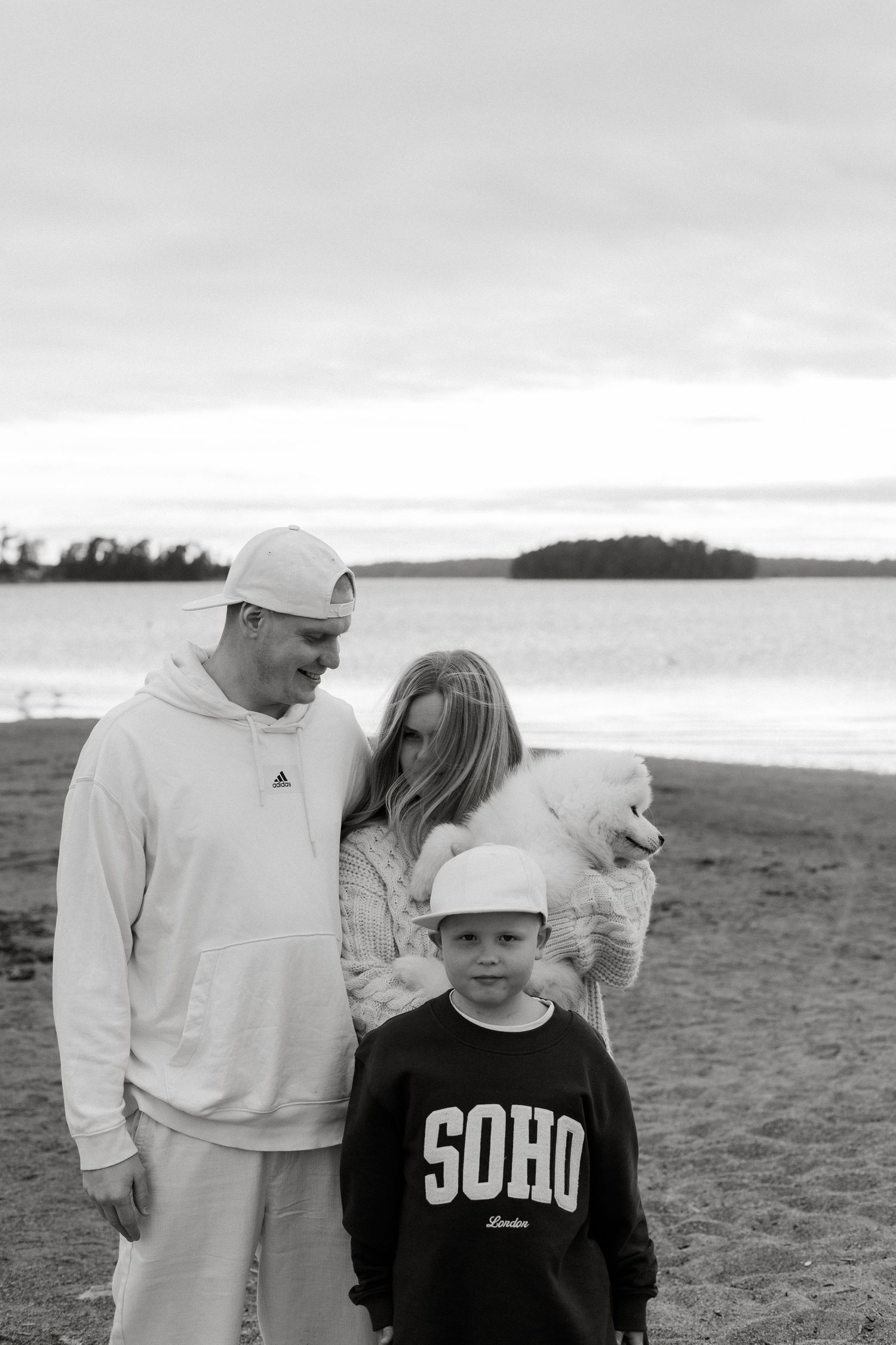 Black and white photo of a family with a young boy in a SOHO sweatshirt posing on a beach with their dog.