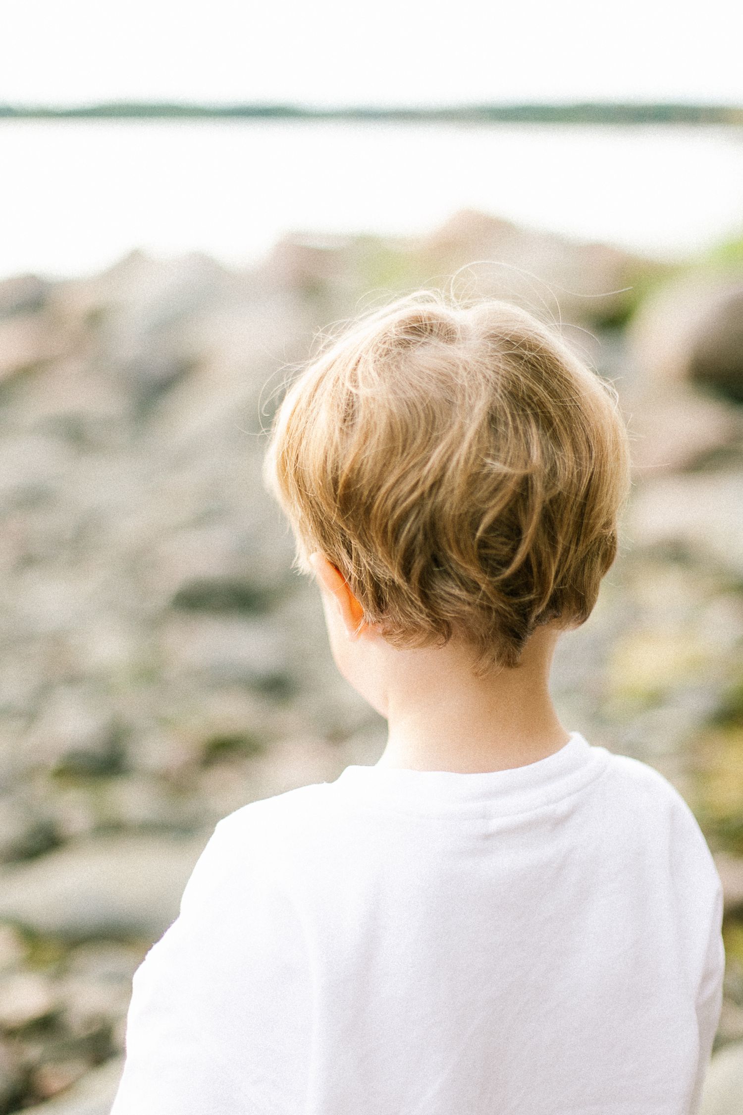 Young blonde child in white shirt viewed from behind, gazing at rocky shoreline and calm water.