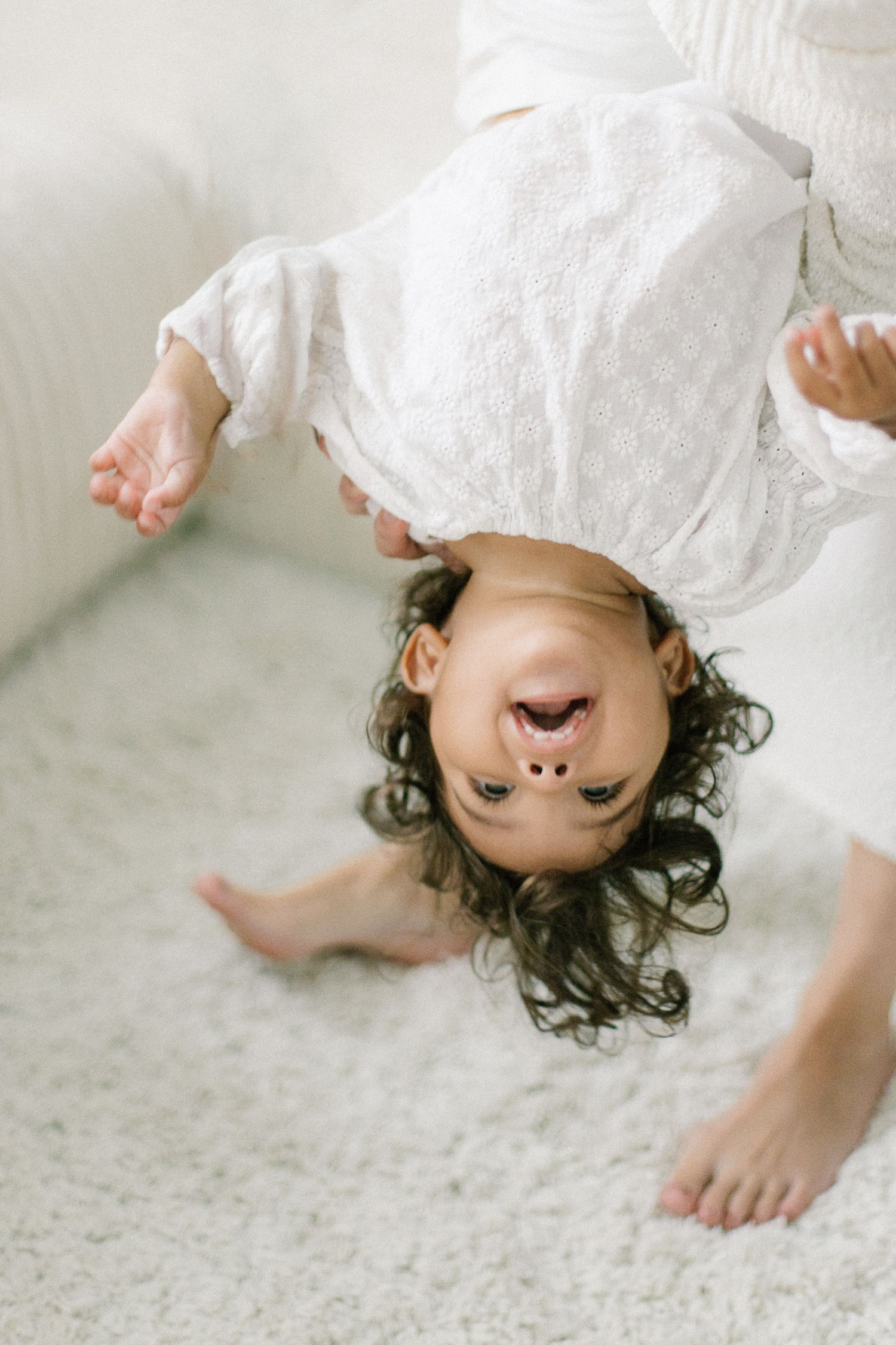 Laughing toddler girl in white dress held upside down, playing on a soft white carpet.