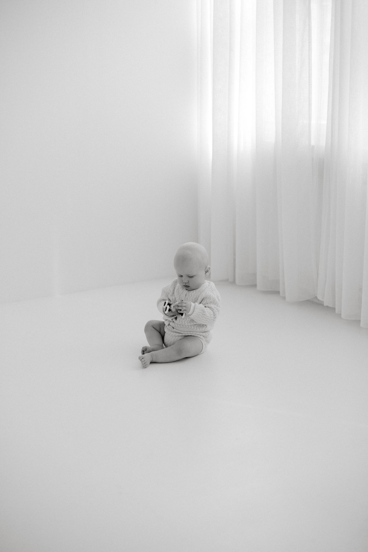 Black and white photo of a baby sitting on a white floor, looking down at a small toy near sheer curtains.