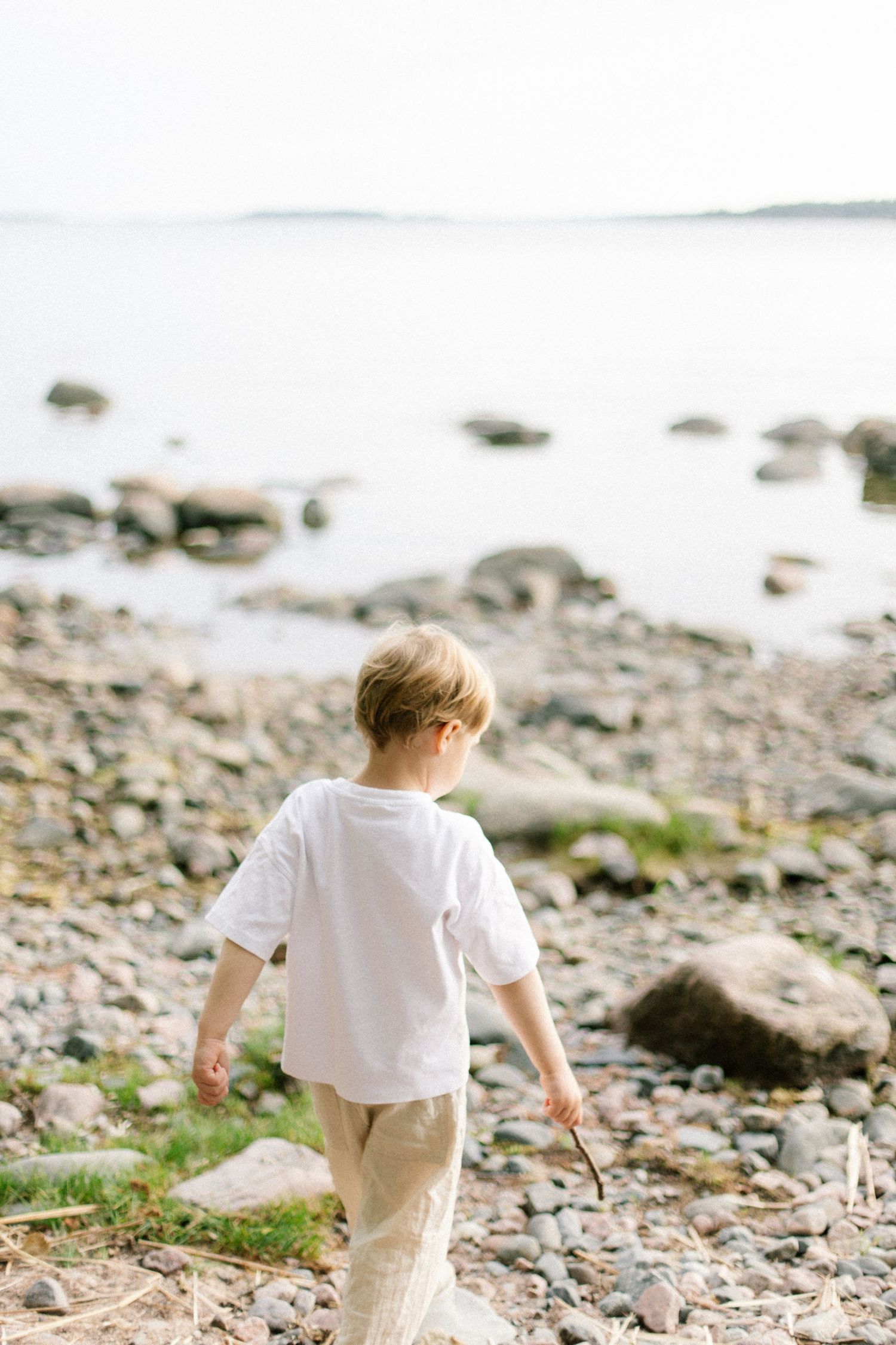 Young boy in white shirt walking on rocky shoreline toward calm ocean water on a clear day.