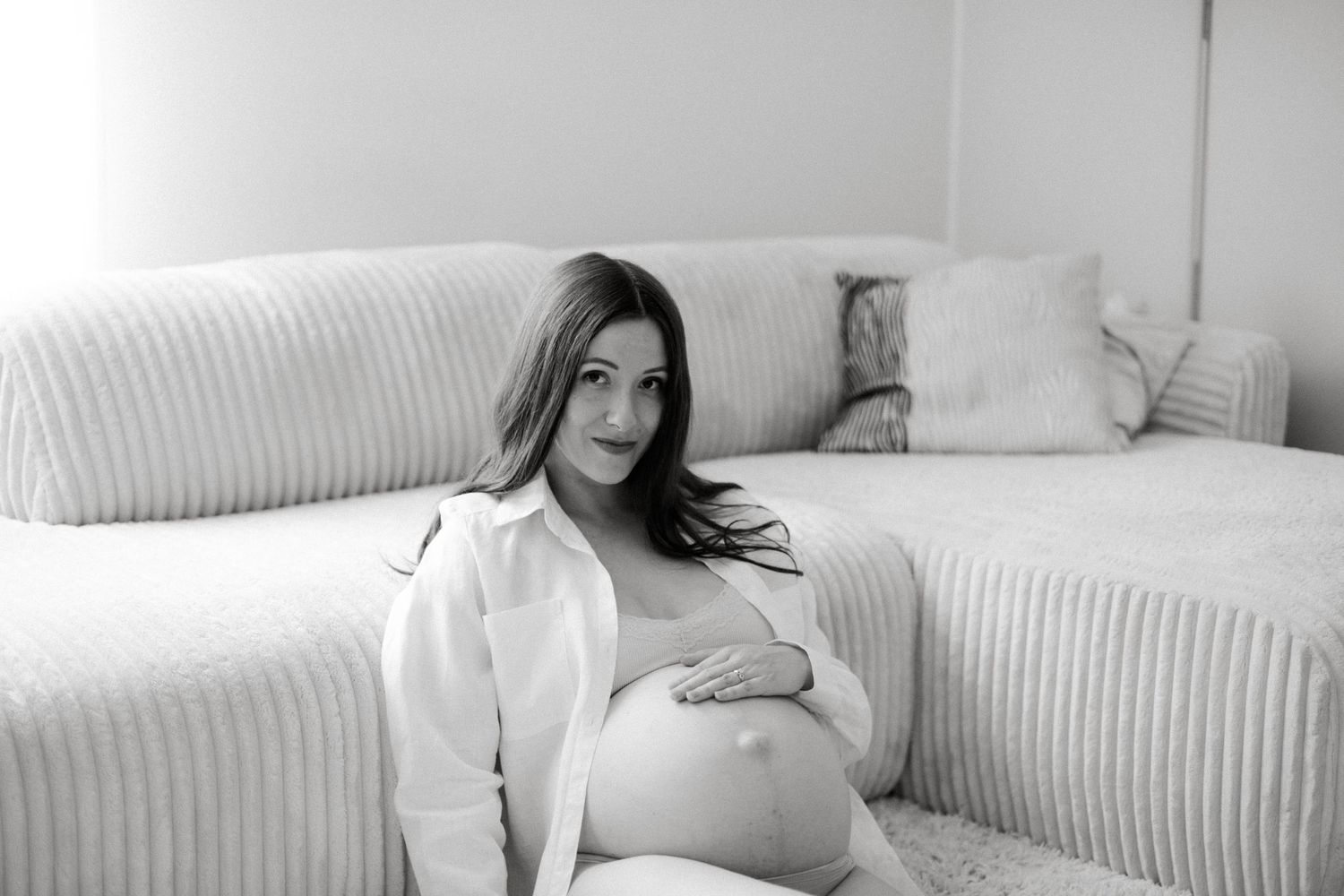 Pregnant woman in white shirt sitting on floor leaning against sofa, black and white maternity photo.