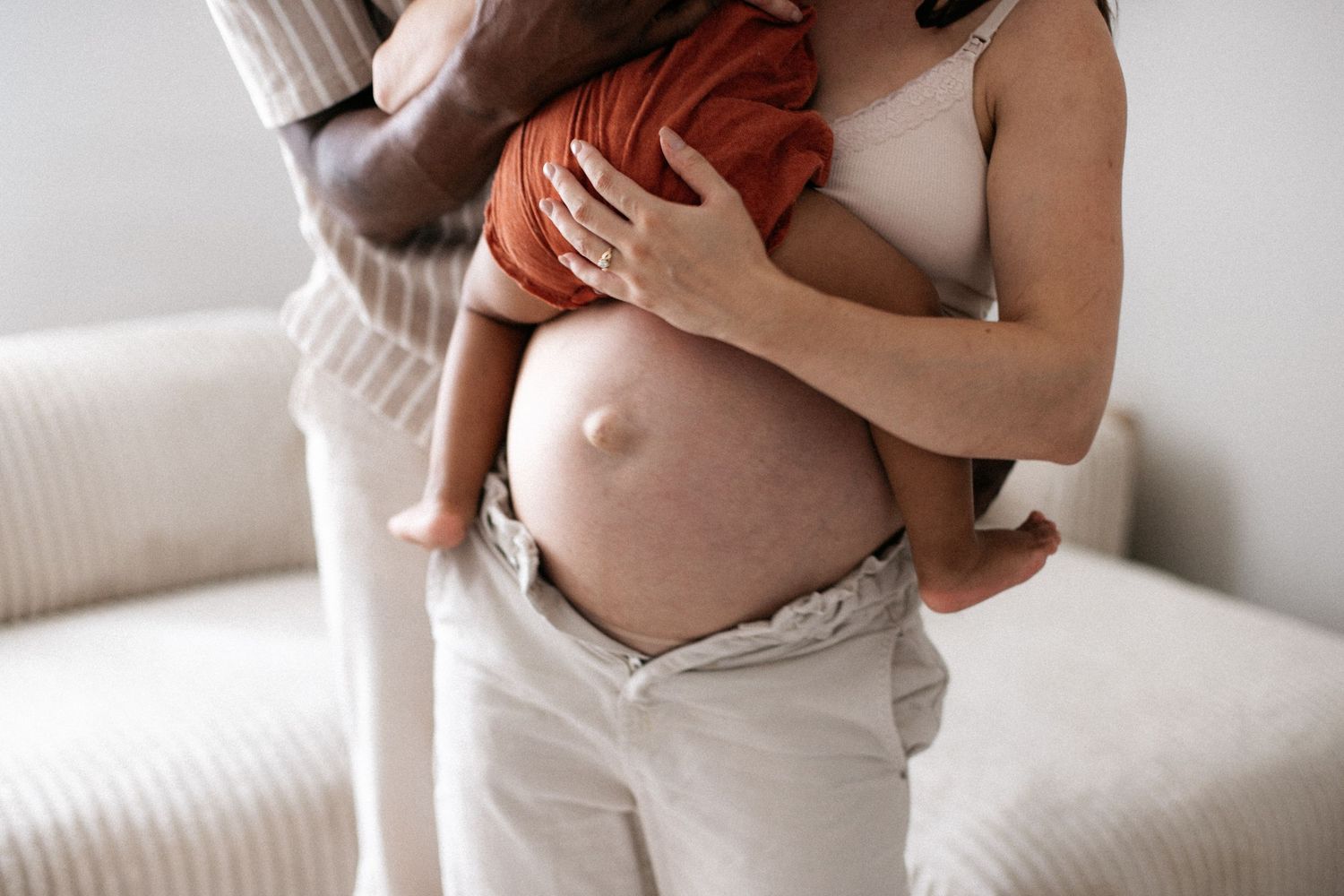 Pregnant woman holding a baby in rust-colored outfit, standing near a beige sofa at home.