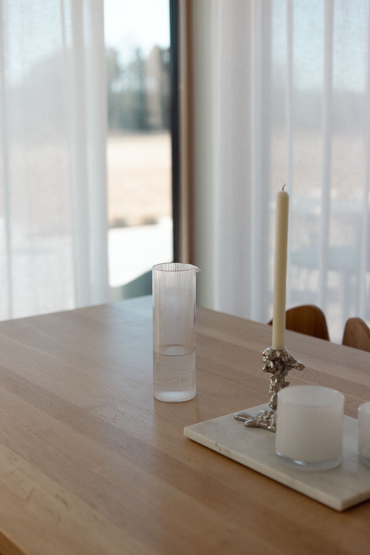 Minimalist wooden dining table with ribbed glass vase, marble tray, silver candleholder, and white pillar candle near sheer curtains.
