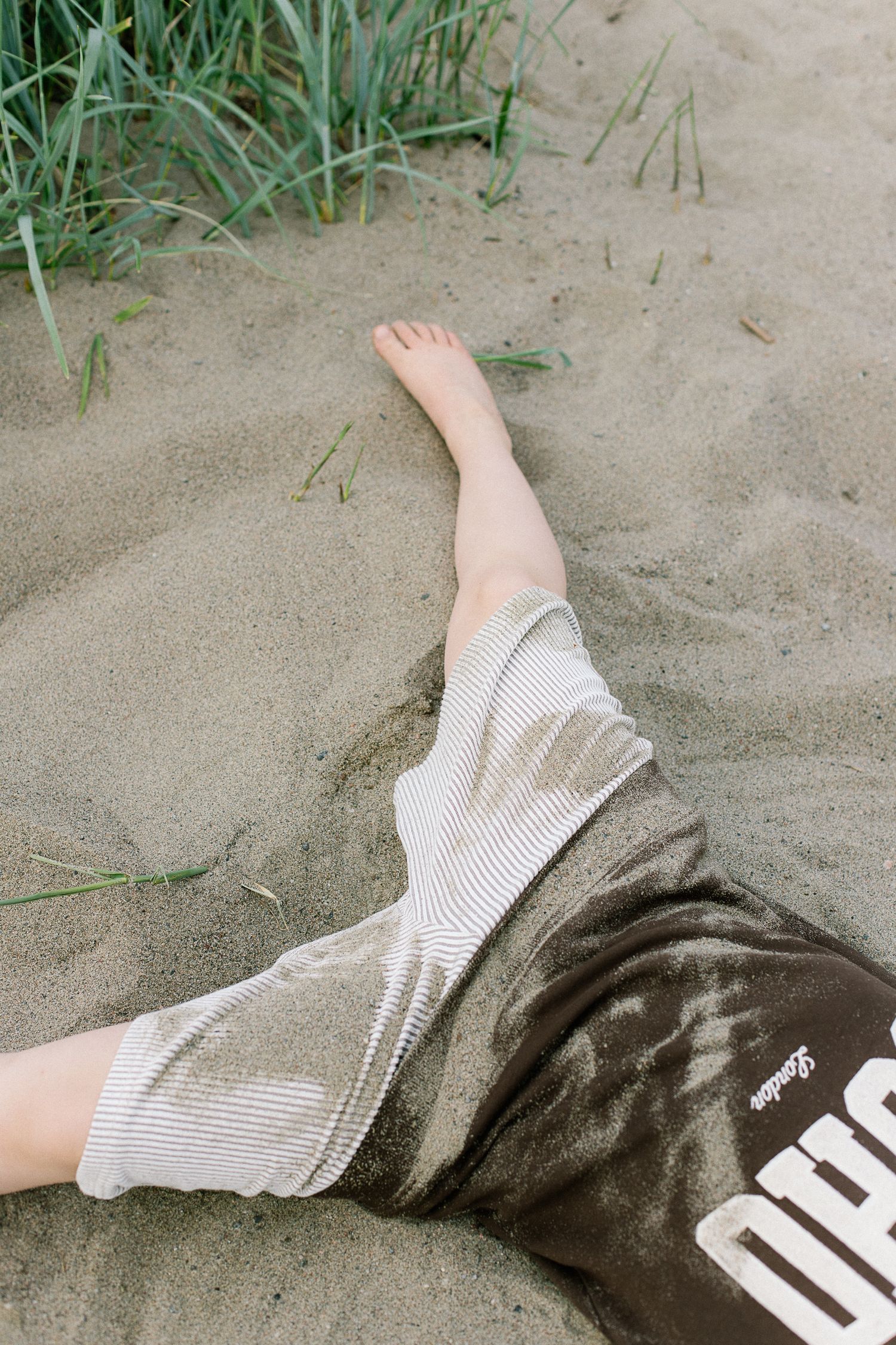 Person relaxing on sandy beach wearing striped shorts and dark graphic shorts with bare feet near green grass.