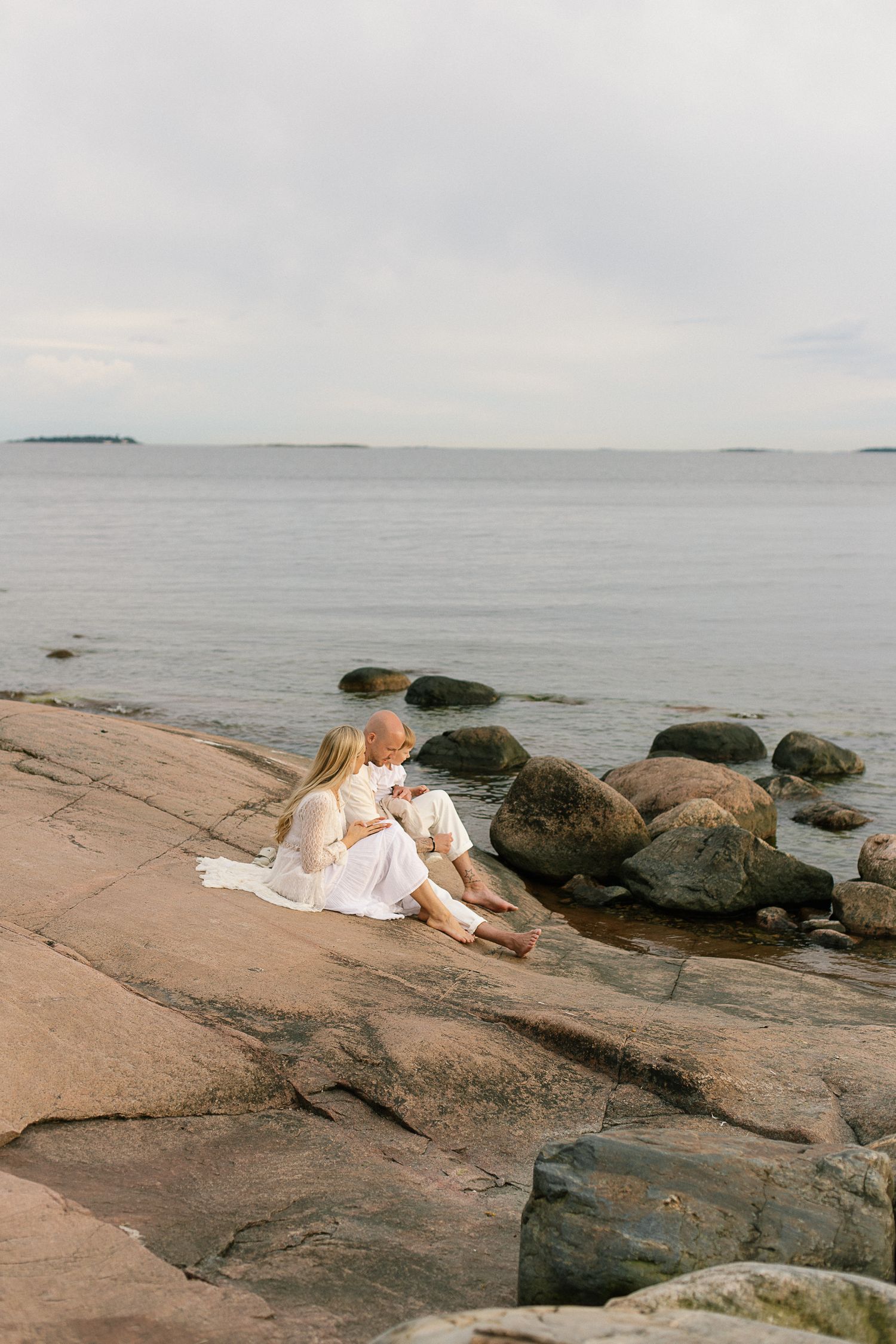 Couple sitting together on rocky coastal shoreline, dressed in white, gazing at calm sea under overcast sky.