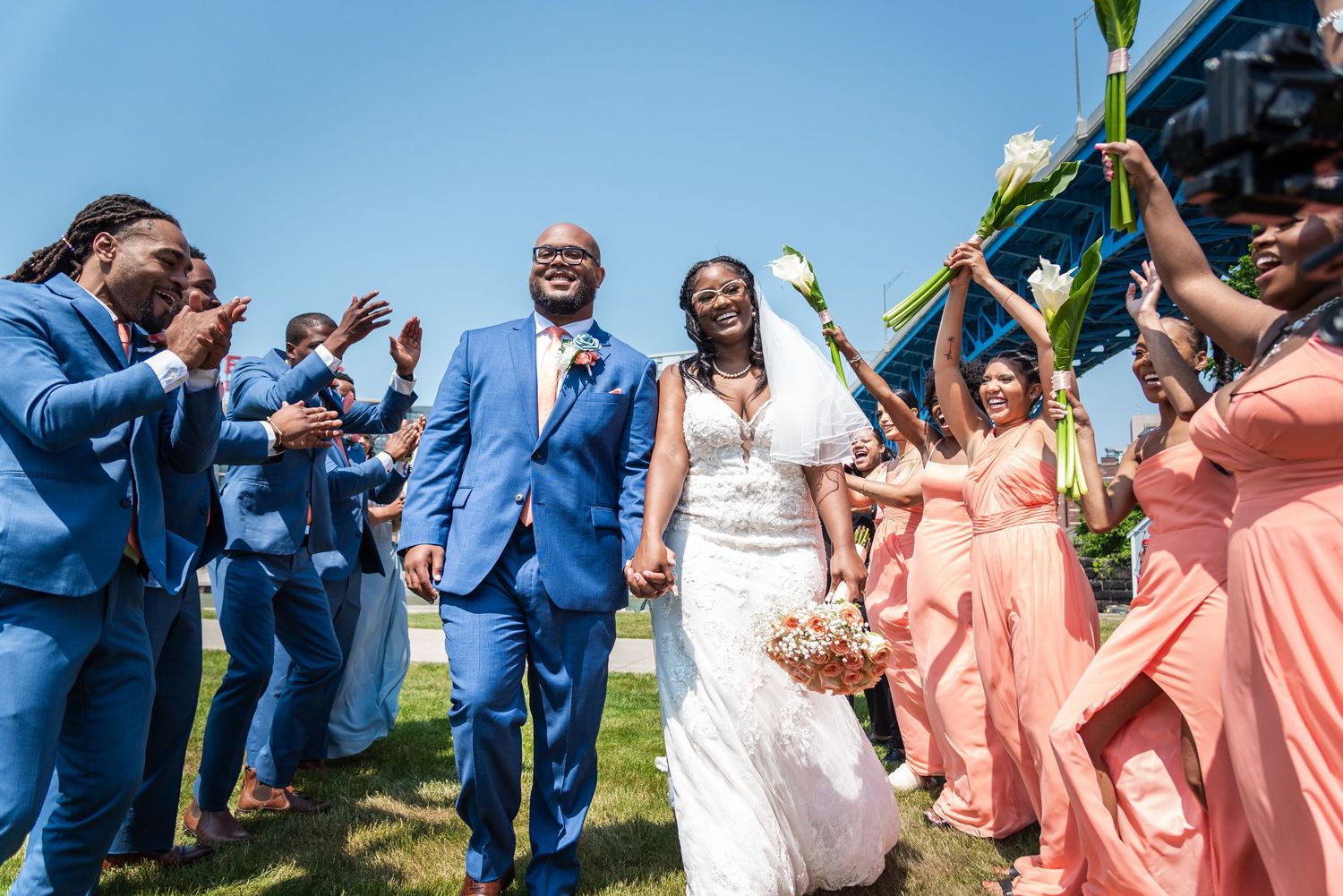 Wedding party celebrates outdoors wearing blue suits and coral dresses.