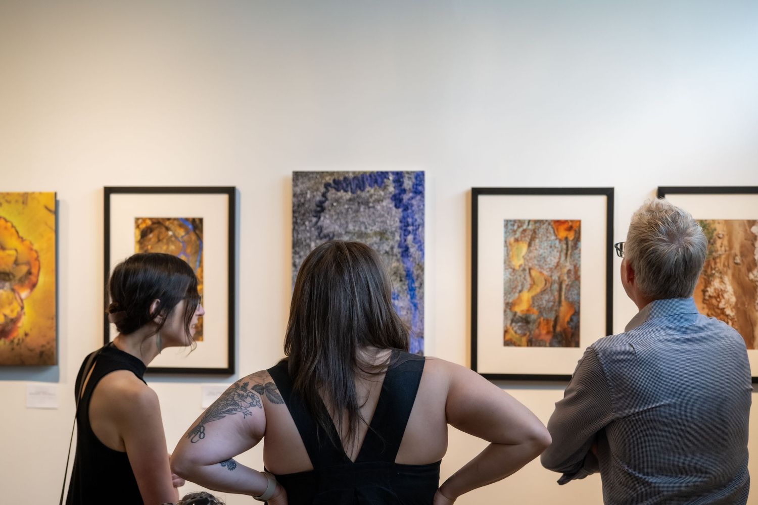 Three visitors viewing framed abstract paintings on a white gallery wall during an art exhibition.