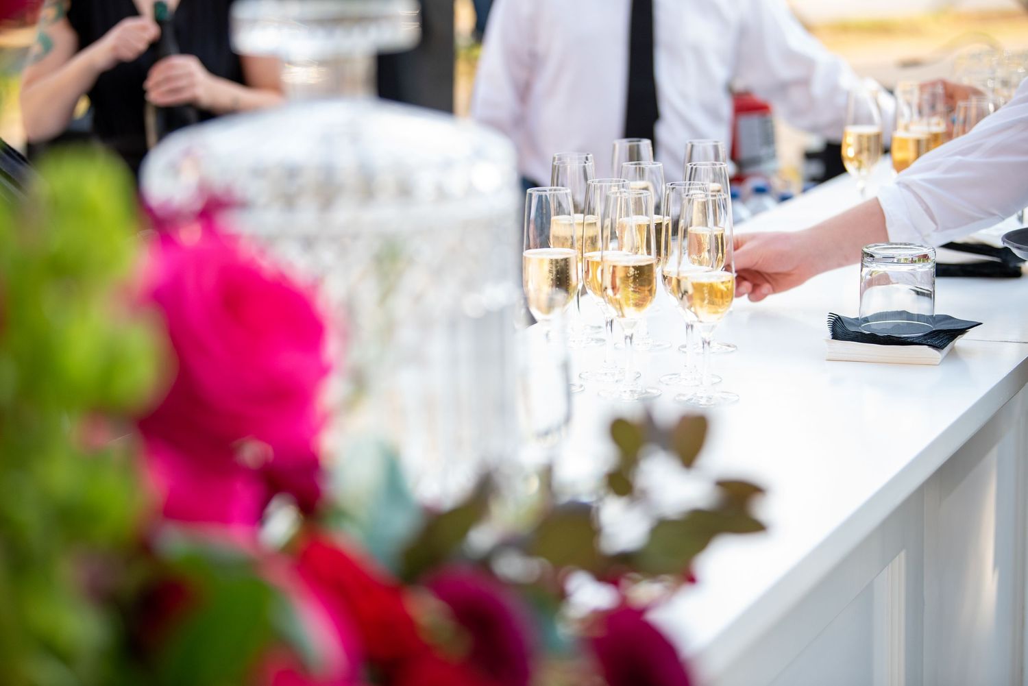 Champagne glasses on a white bar counter at an elegant outdoor event with pink floral decorations.
