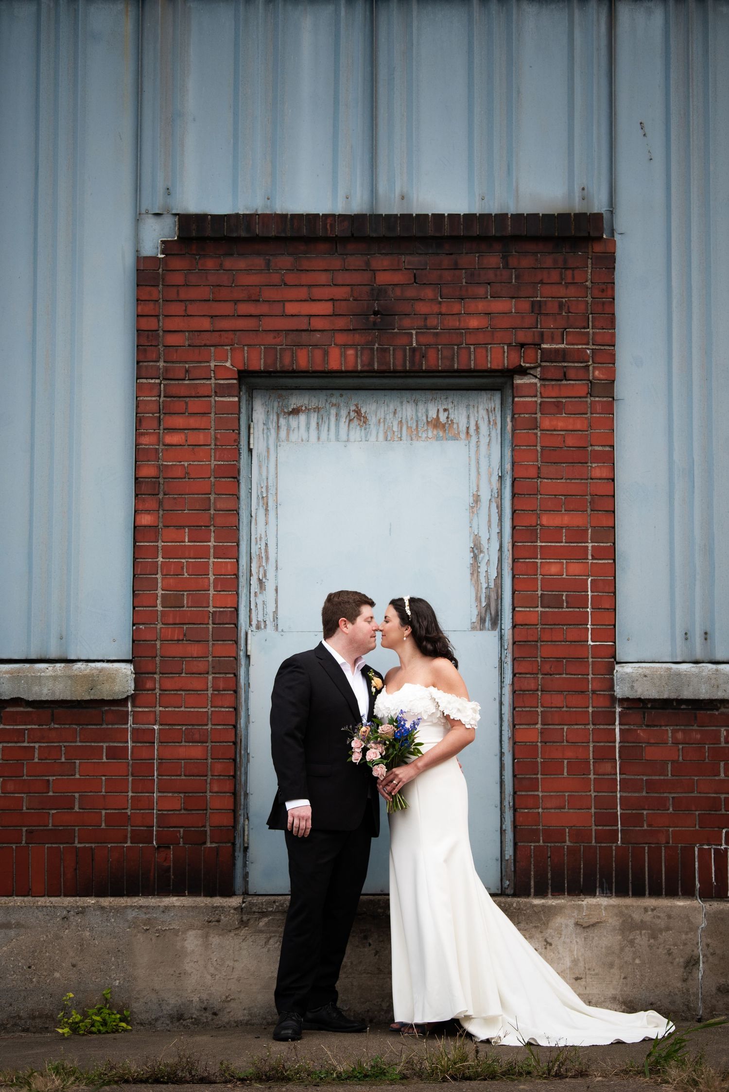 Wedding couple poses against rustic red brick wall with white industrial doors.