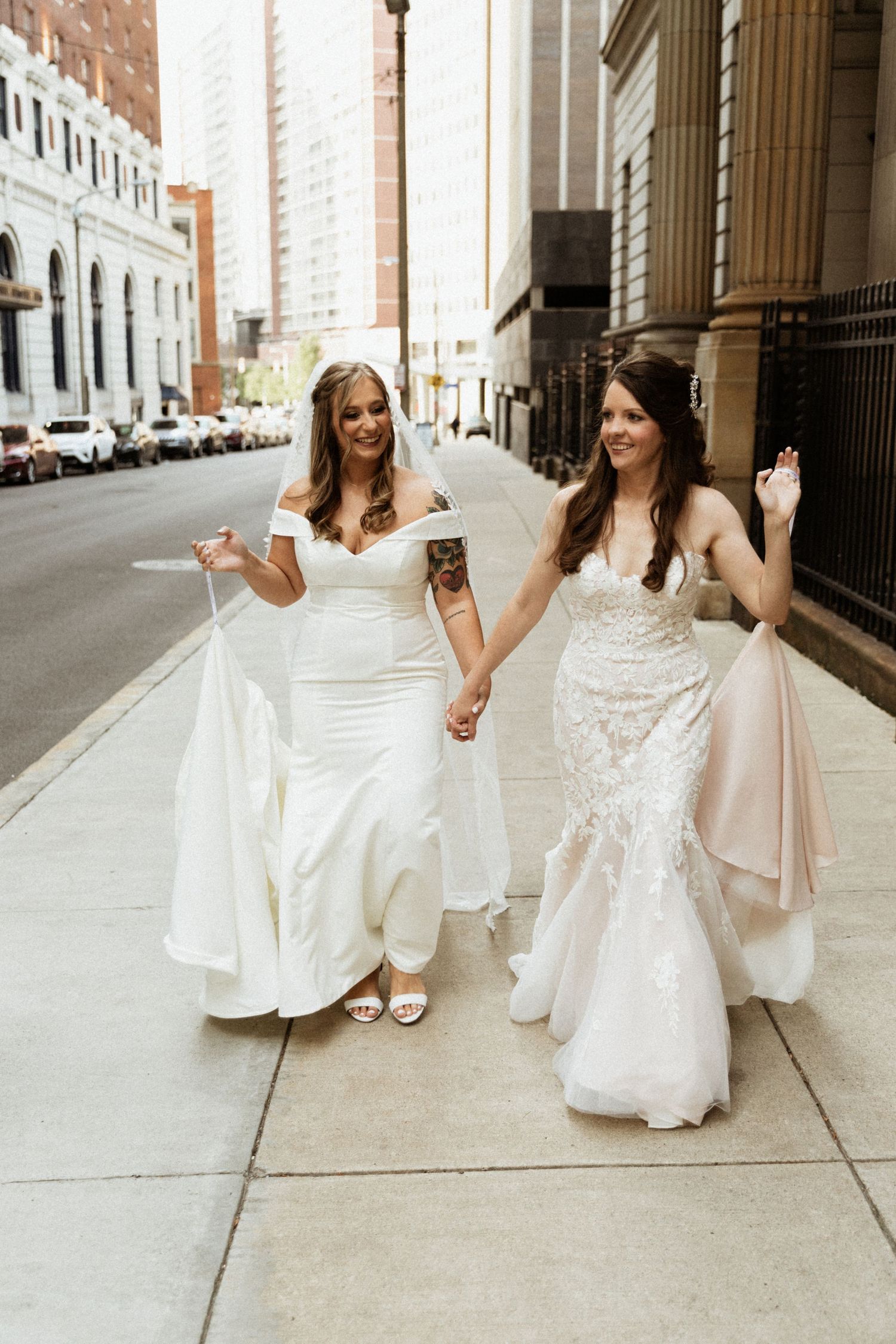 Two brides in white wedding gowns hold hands while walking down a city sidewalk, smiling joyfully on their wedding day.