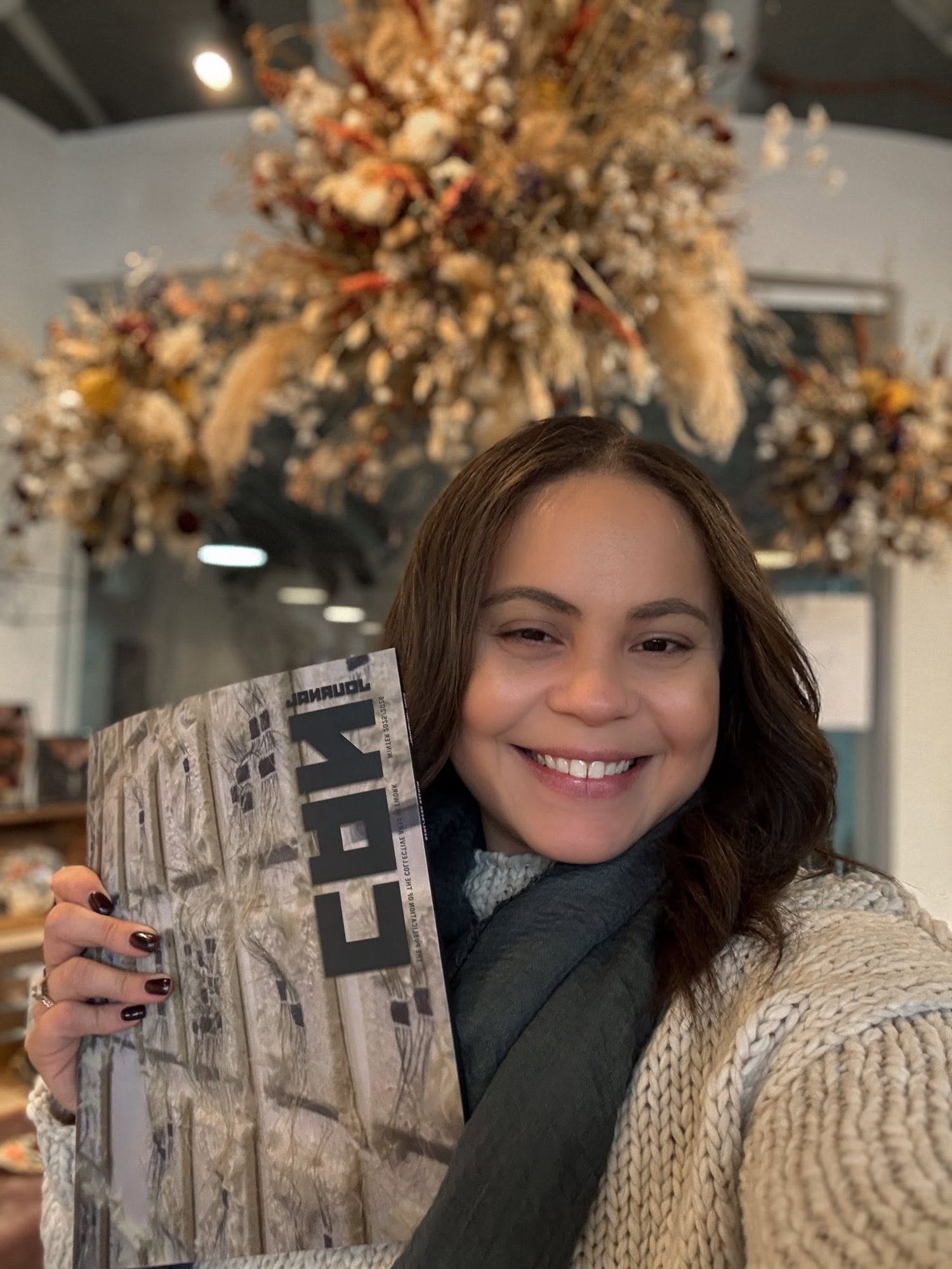 Smiling woman holding a magazine in a cozy shop with dried floral arrangements overhead.
