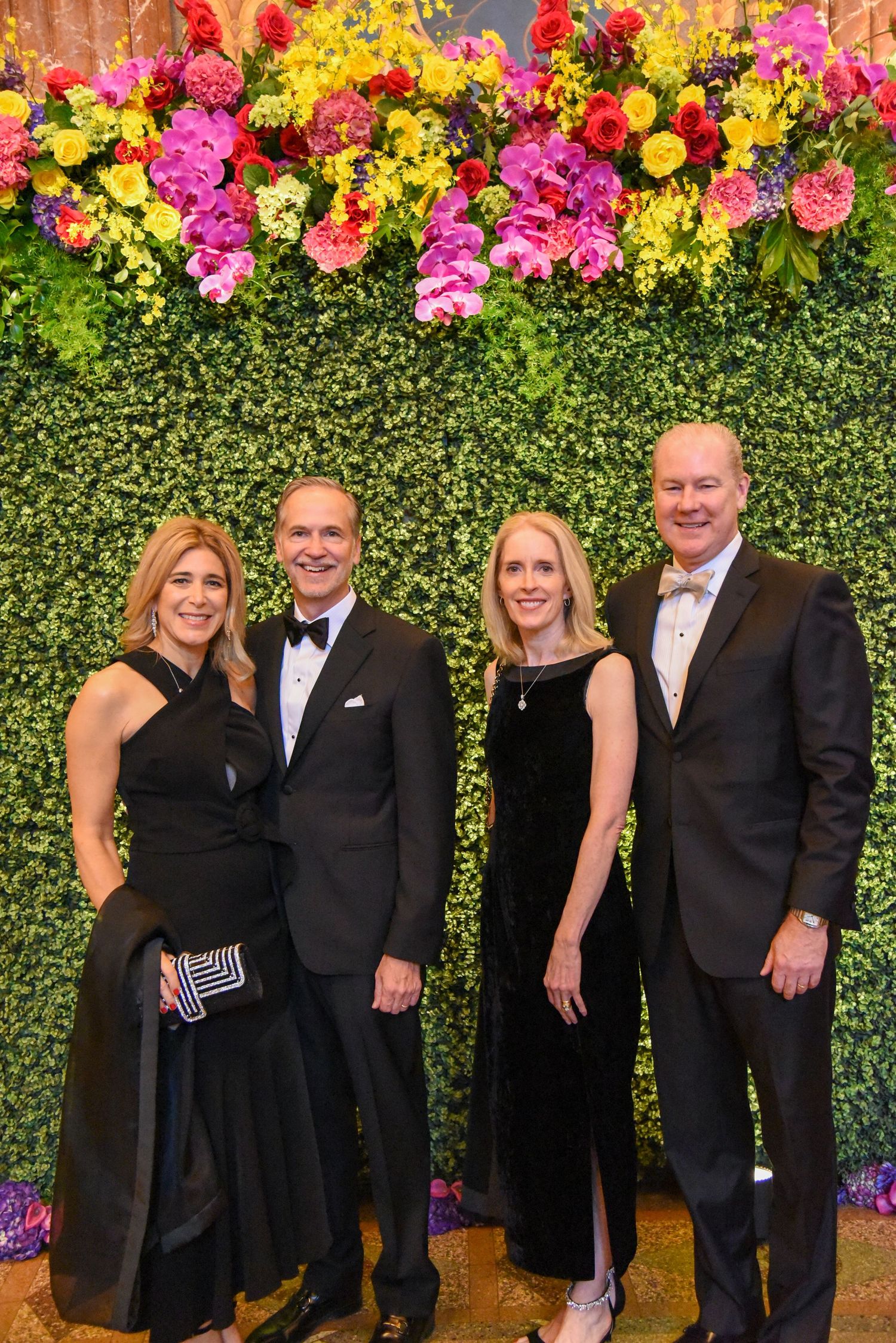 Formal event photo backdrop featuring vibrant pink, yellow and red flowers against a hedge wall.