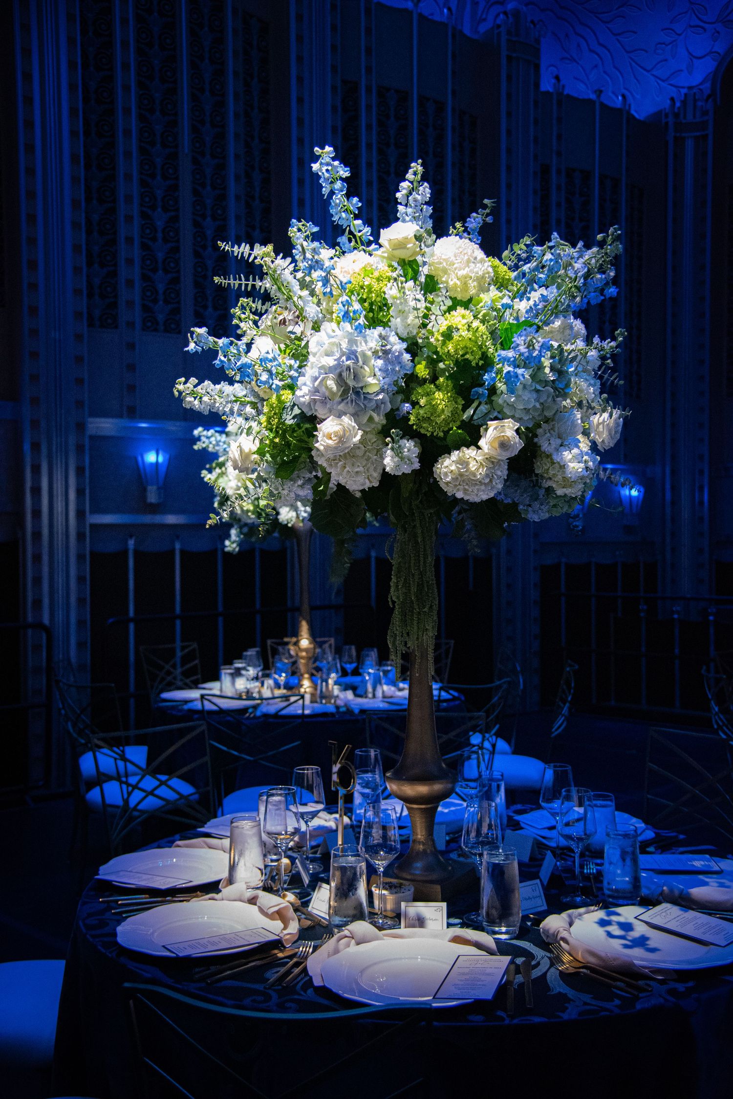 Dramatic blue-lit reception tables feature tall white floral arrangements and formal place settings.