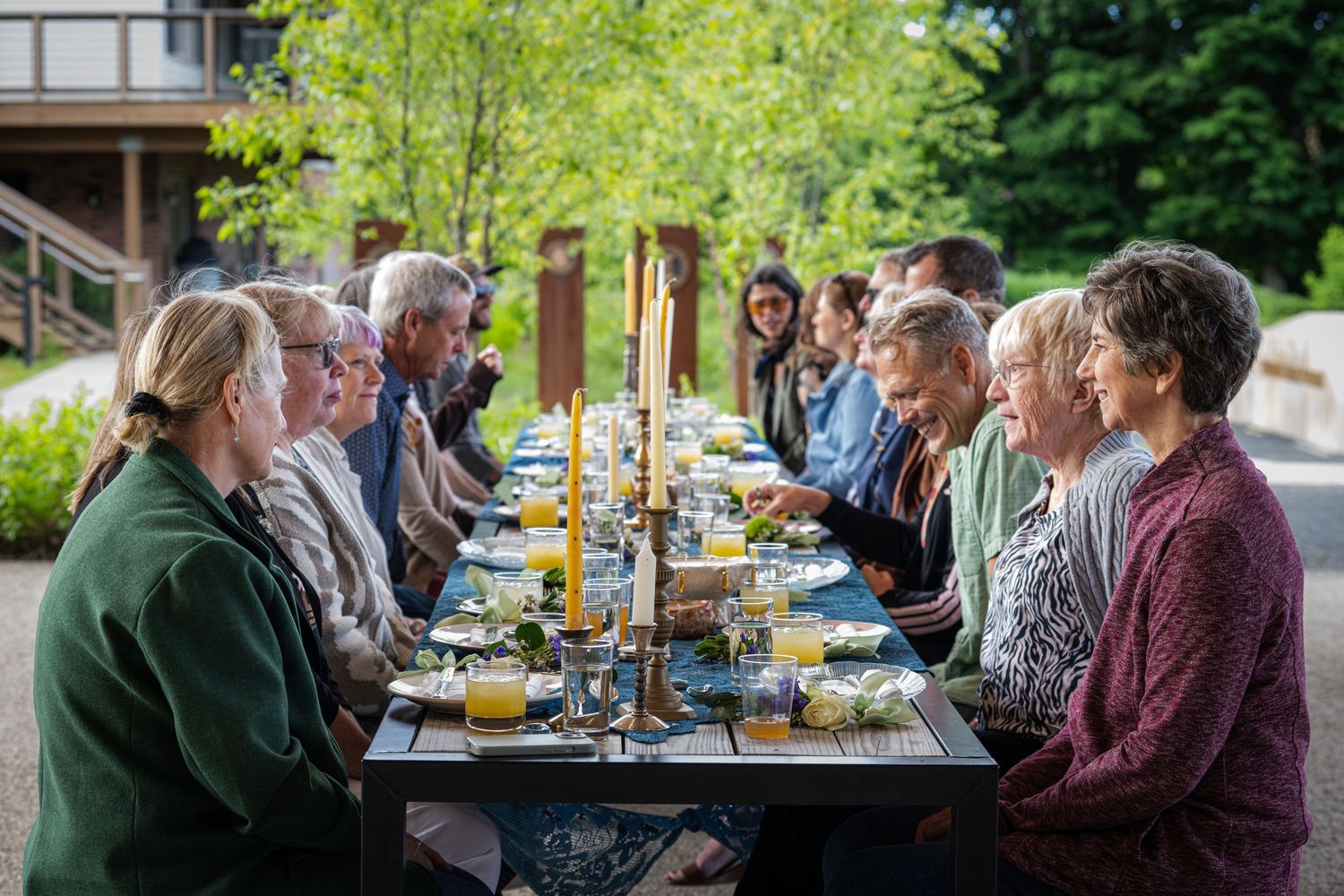 Group of people dining outdoors at a long table surrounded by lush green trees during a garden party.