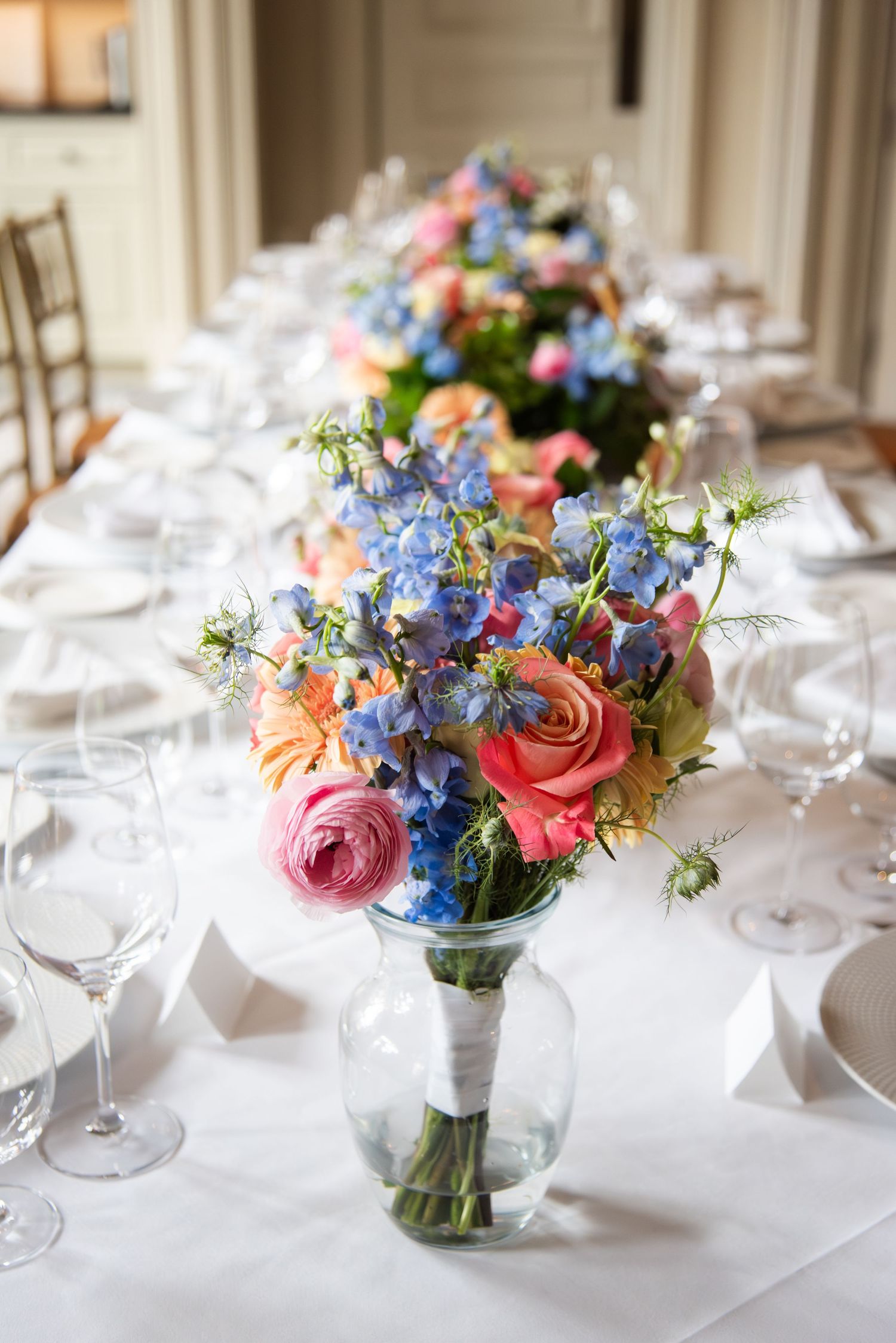 Colorful floral centerpieces with pink roses and blue delphinium arranged on elegant white tablescapes.