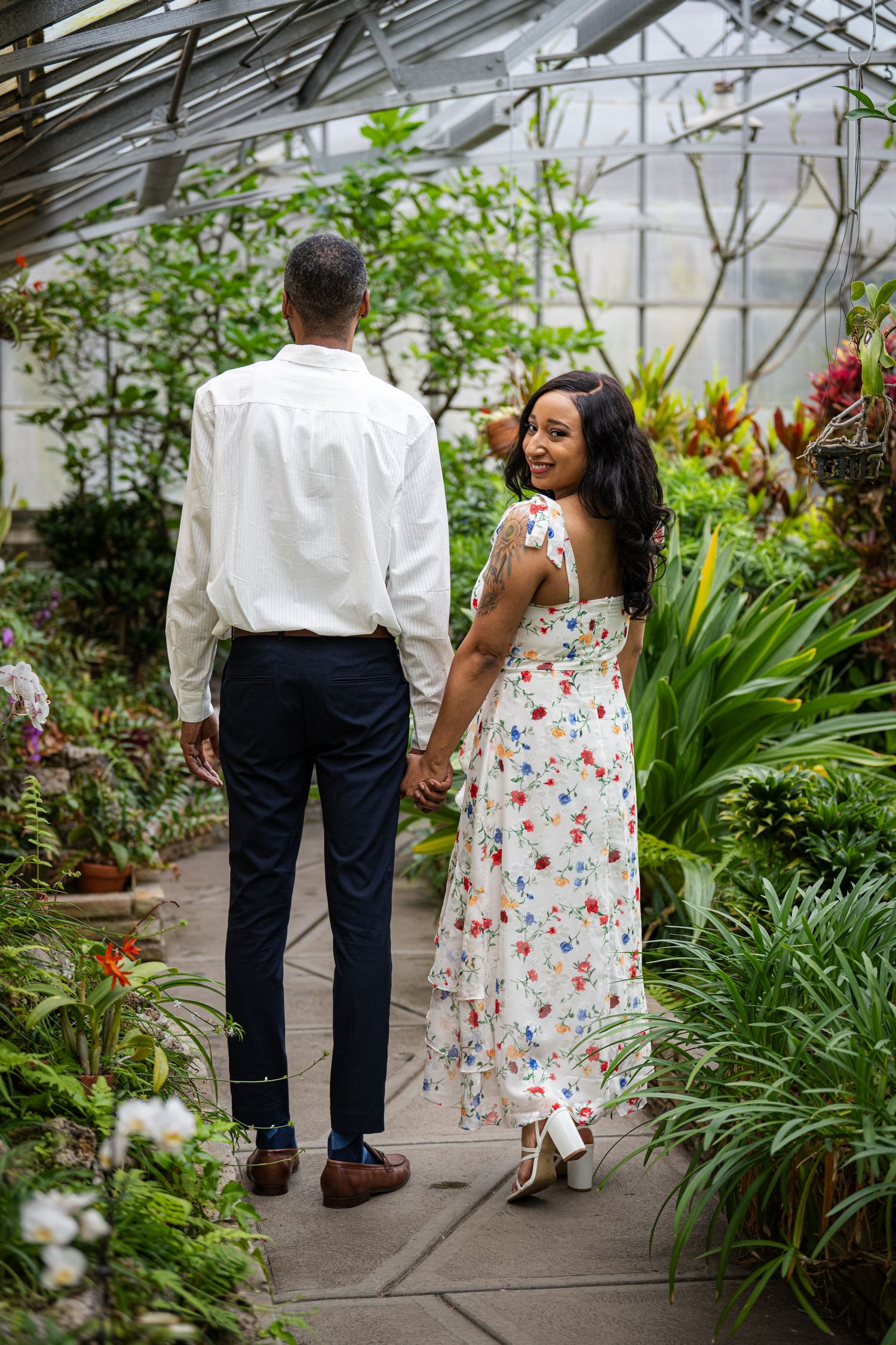 A couple walks through a greenhouse pathway lined with tropical plants and flowers.