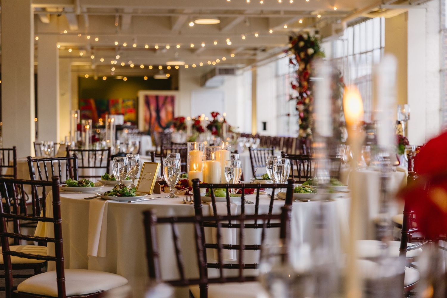 Elegant wedding reception venue with string lights, chiavari chairs, candlelit tables, and floral centerpieces in a bright open space.