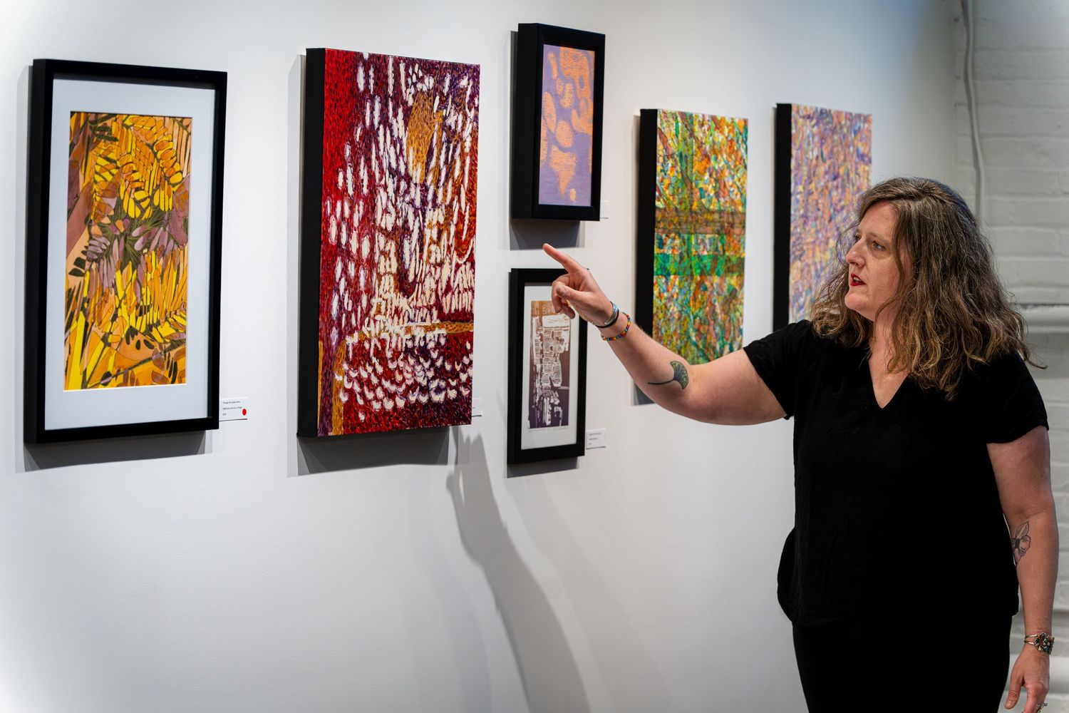 Woman in black outfit pointing at colorful abstract paintings displayed on a white gallery wall.