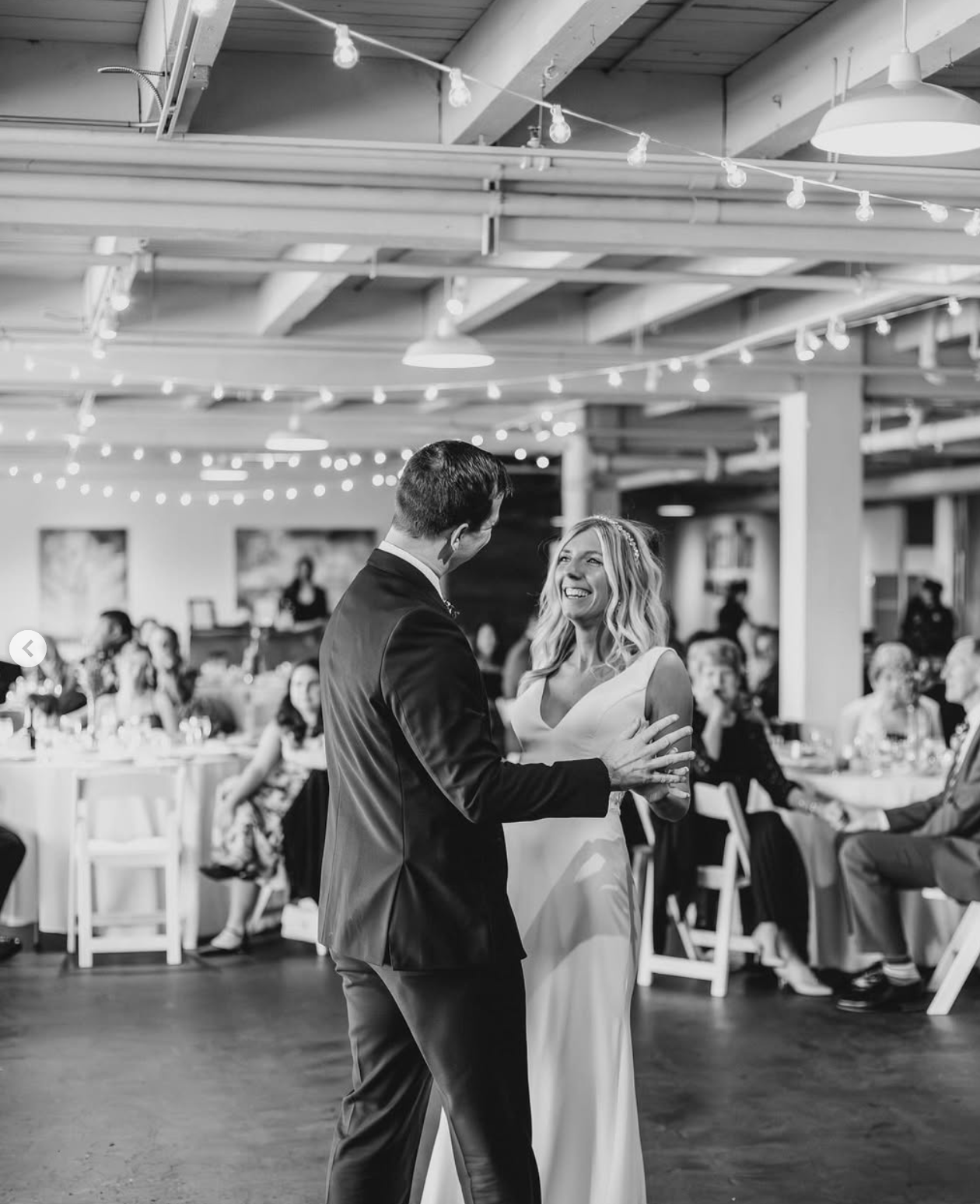 Black and white photo of an indoor reception venue with exposed ceiling beams and string lights.