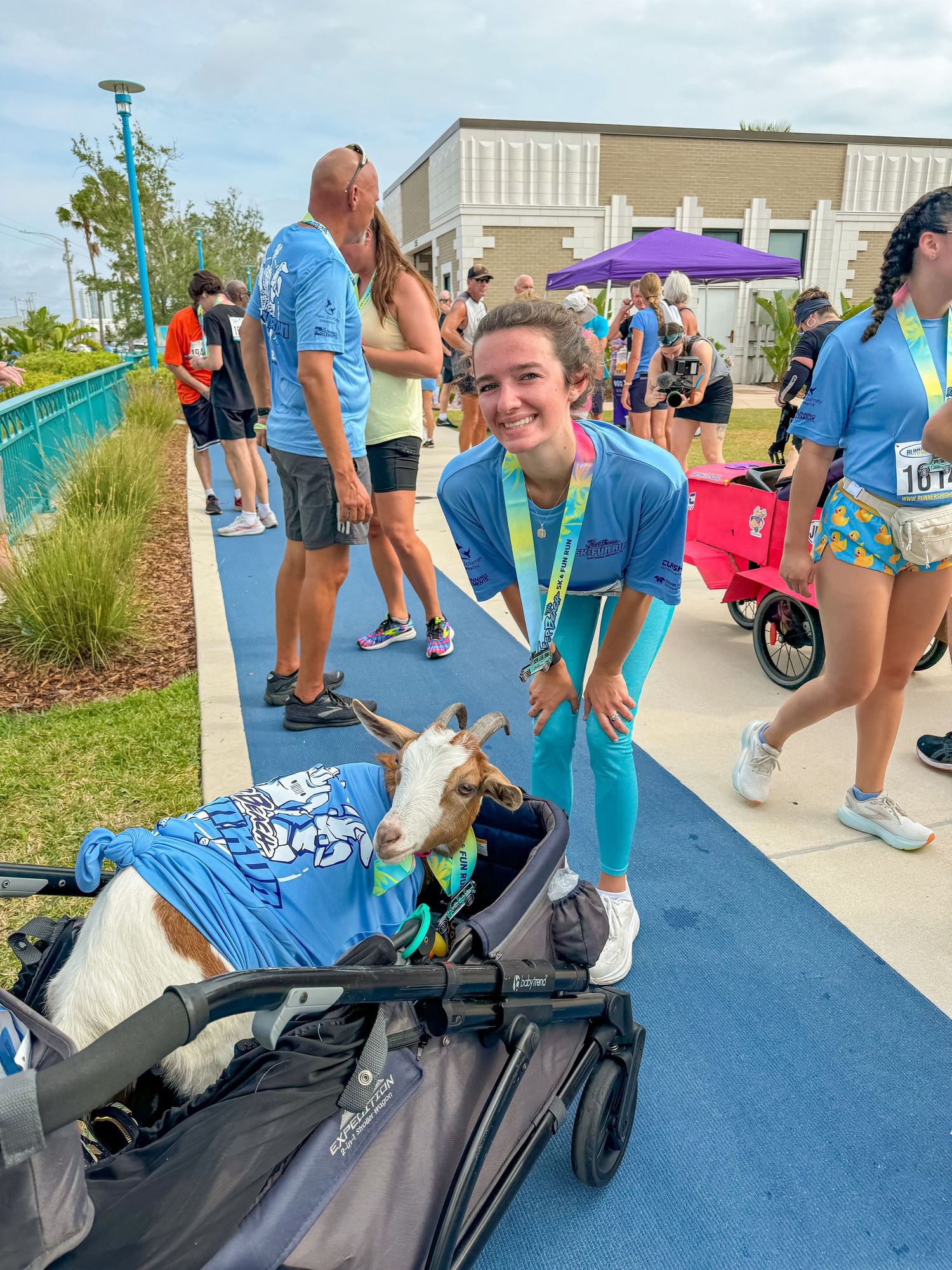 A smiling woman in blue poses with a goat in a stroller at an outdoor event.