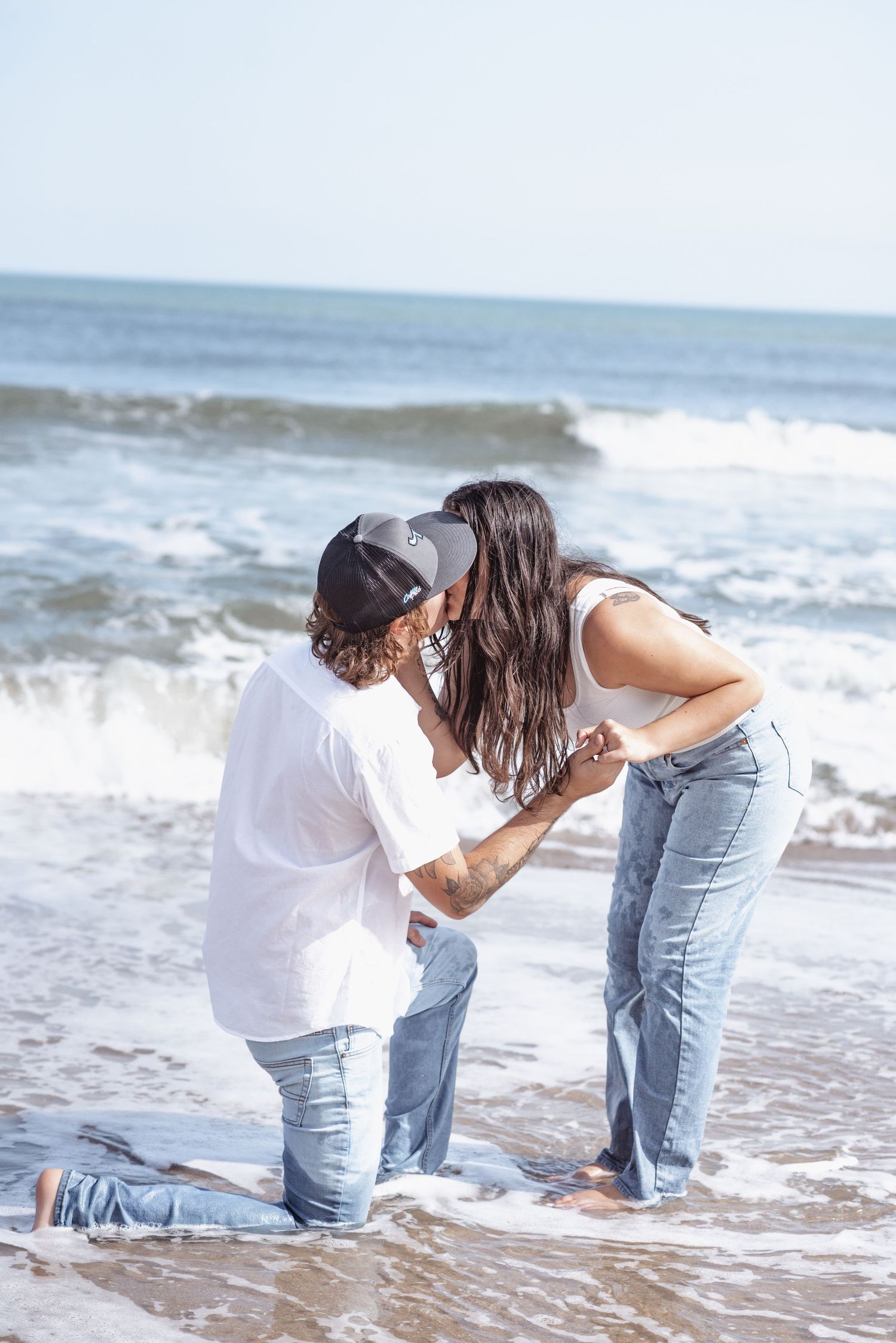 Couple sharing a romantic kiss on the beach, man kneeling in sand while woman stands, both wearing jeans and caps.