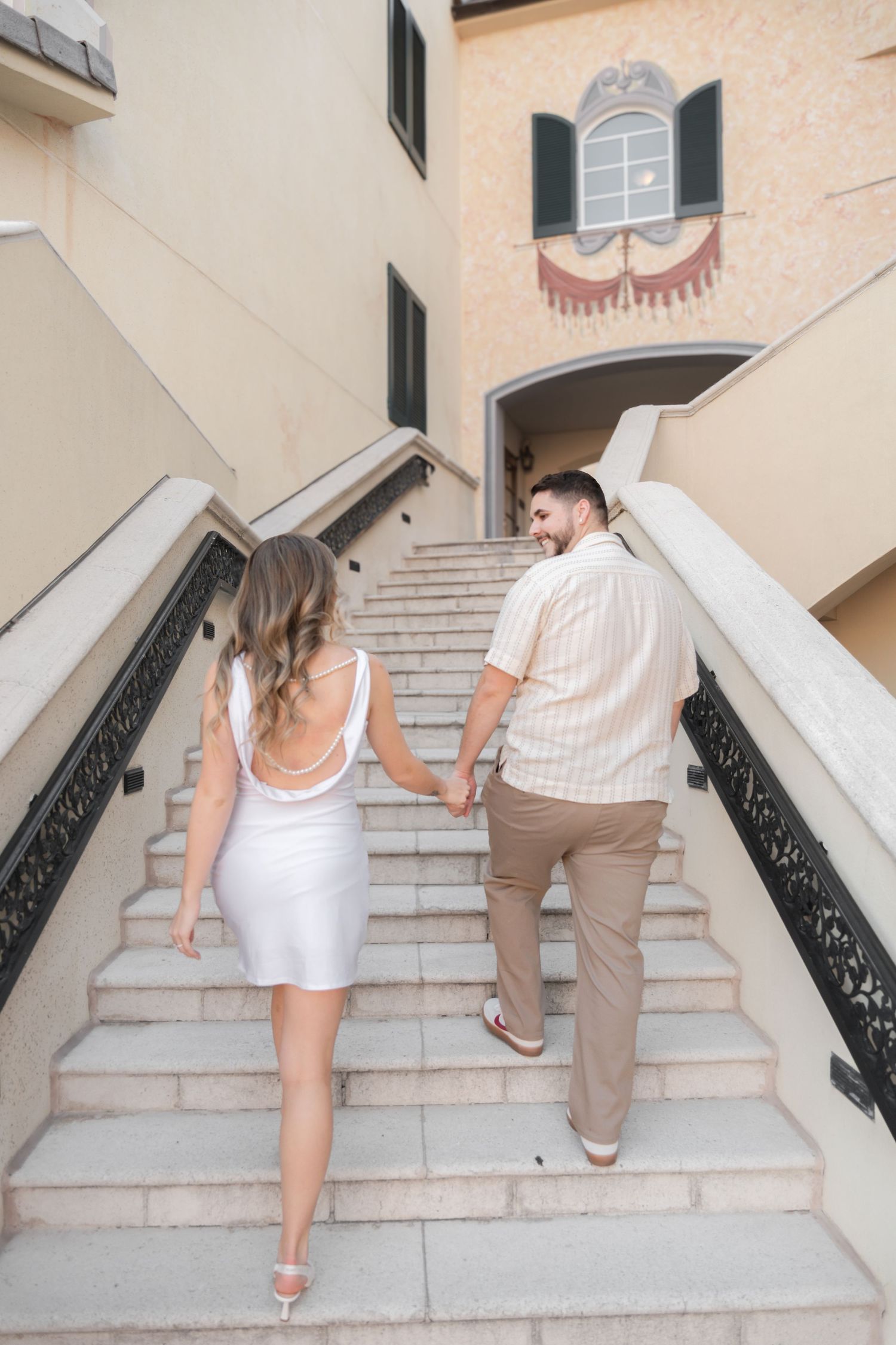 Couple holding hands walking up elegant outdoor stairs, woman in white dress and man in beige shirt.