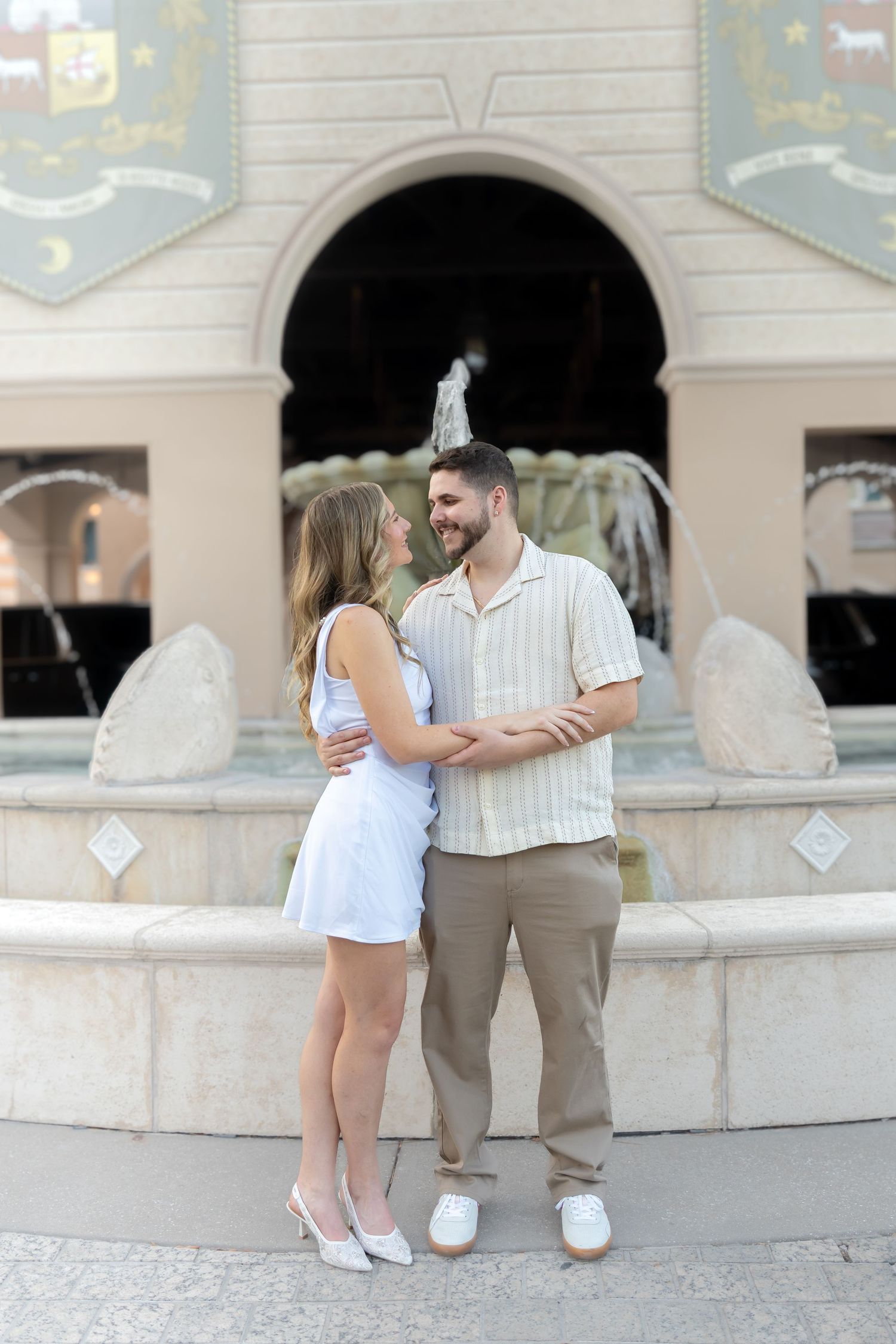 Couple embracing and smiling in front of an ornate fountain at an outdoor plaza.