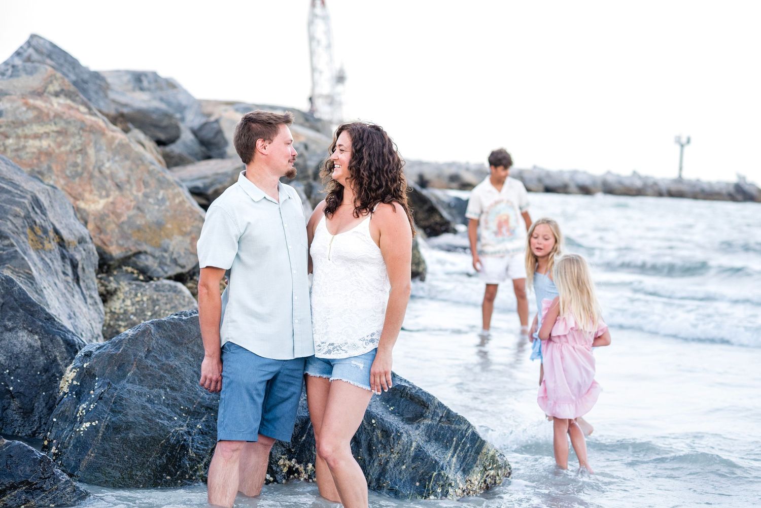 Family enjoying a sunny beach day near rocky shore, couple smiling at each other with children playing in ocean waves.