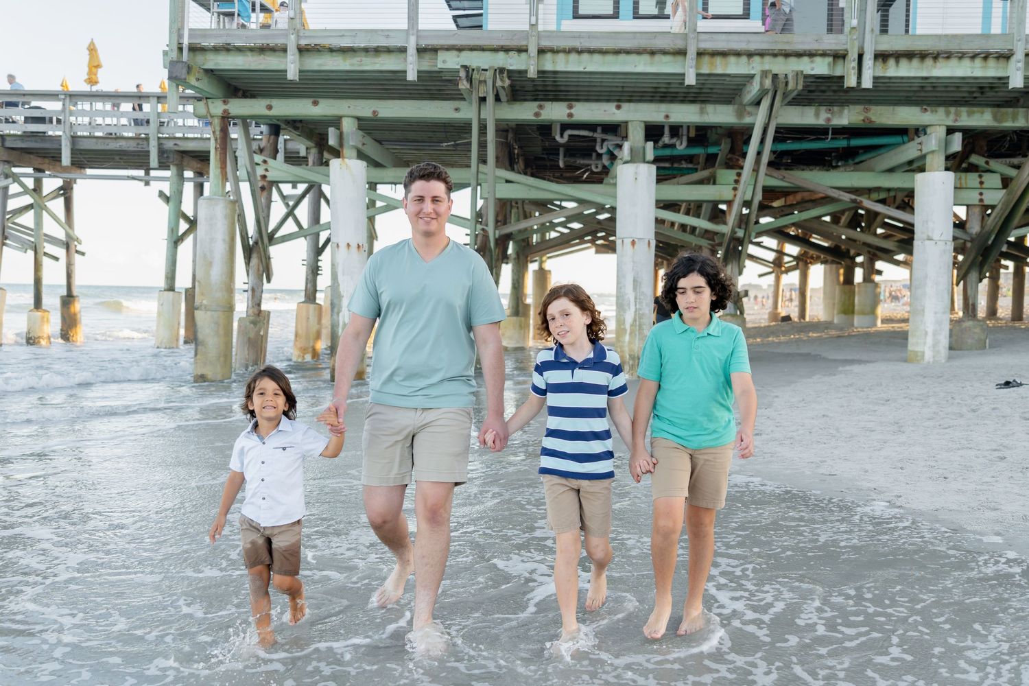 A family of four walks along a beach near a pier, with waves at their feet on a sunny day.