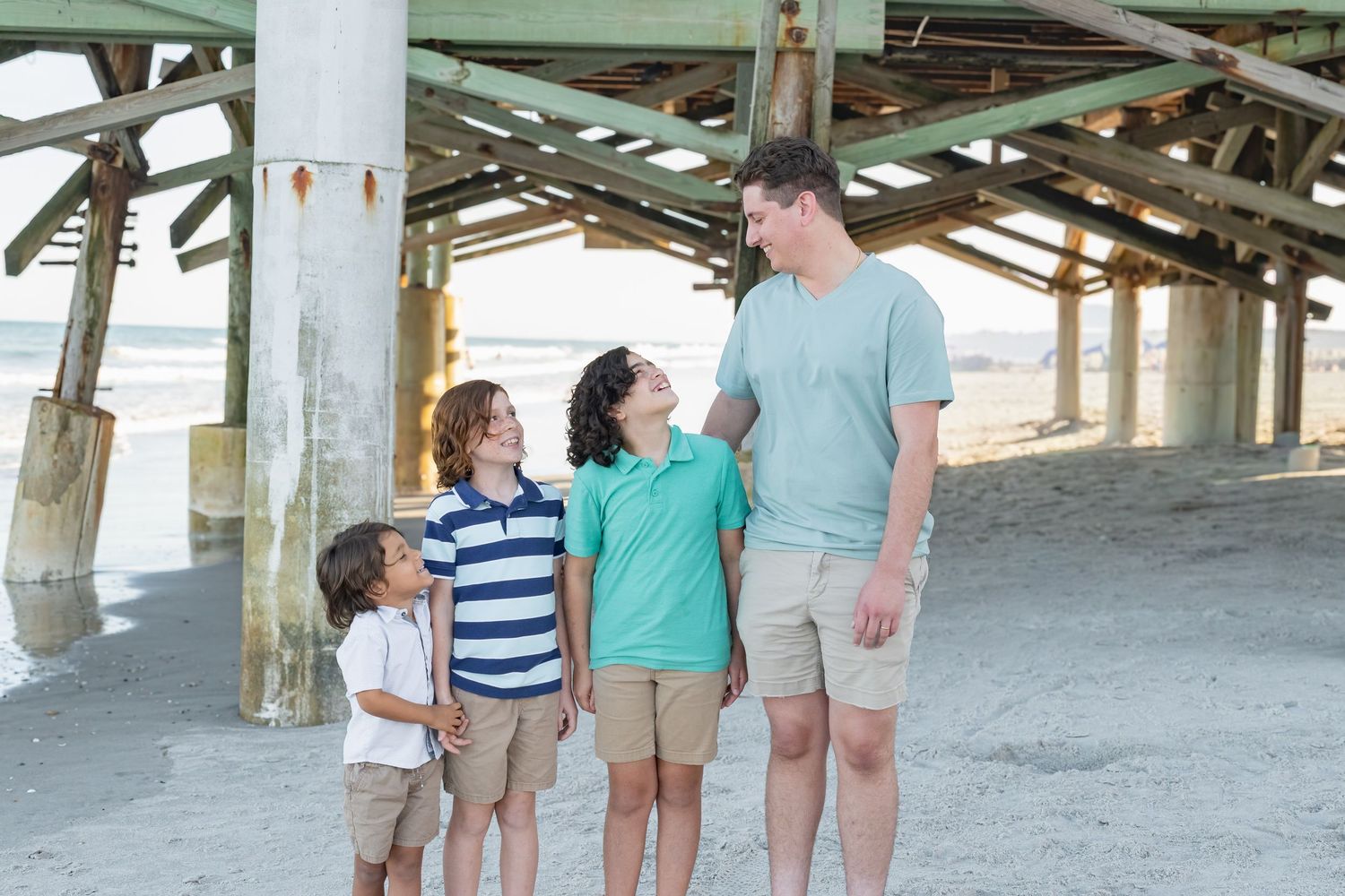 A father and three sons smile and interact under a wooden beach pier on a sunny day.
