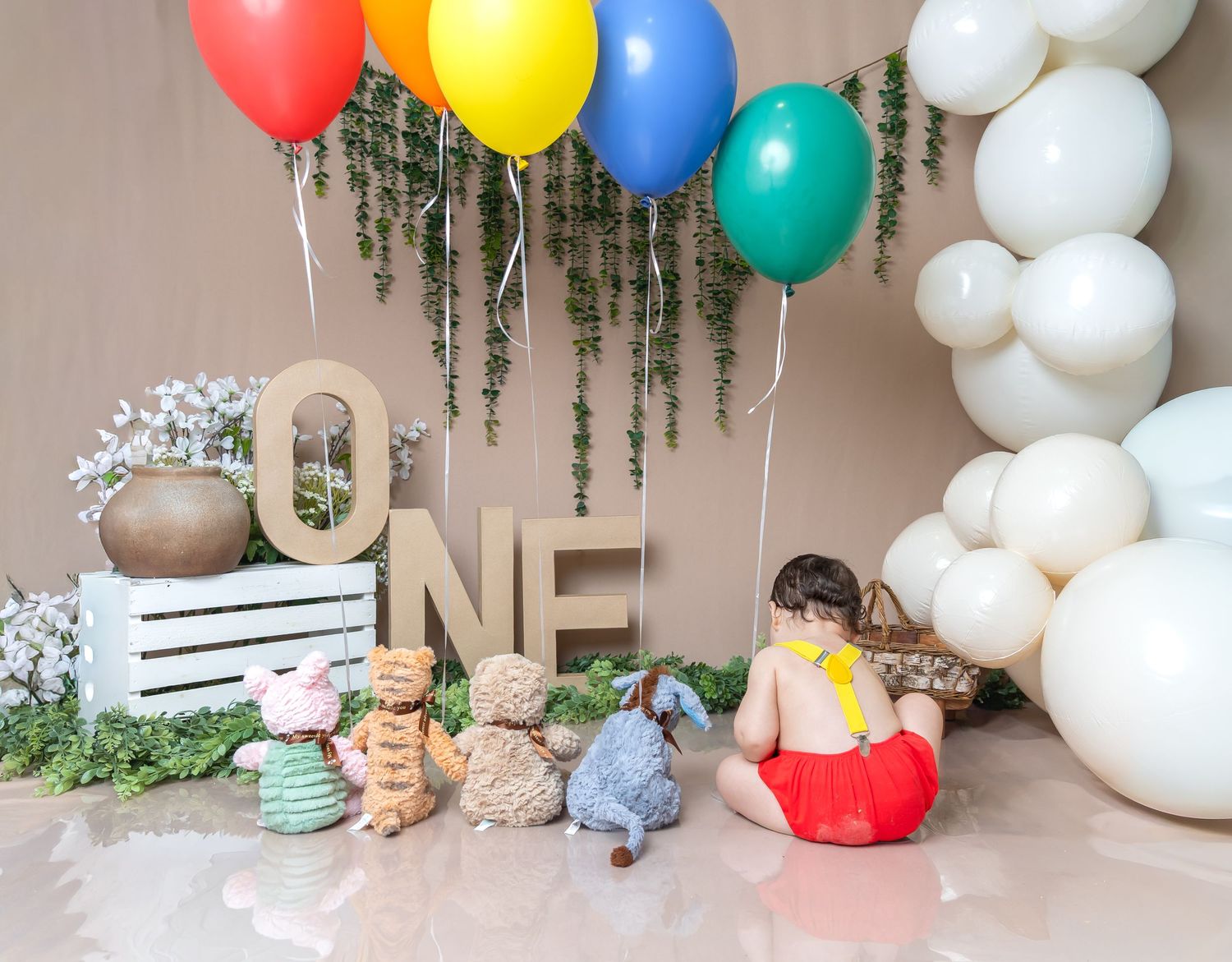 One-year-old baby sitting before colorful balloon arch, ONE sign, and Winnie the Pooh stuffed animals display.