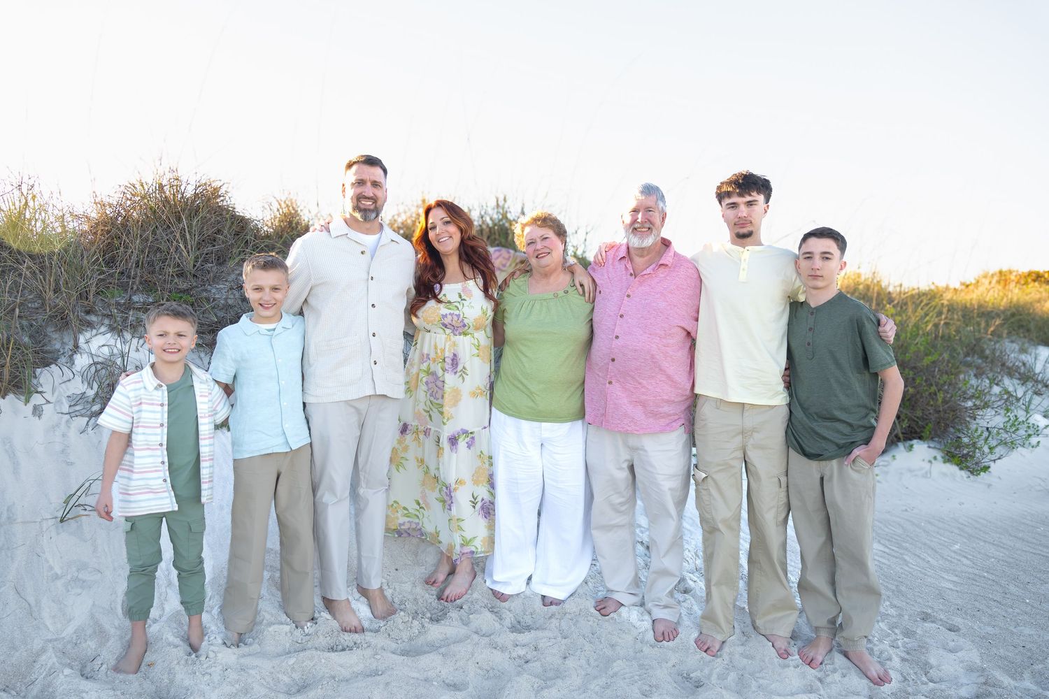 Family of eight poses together on a sandy beach at sunset, dressed in coordinating pastel outfits.