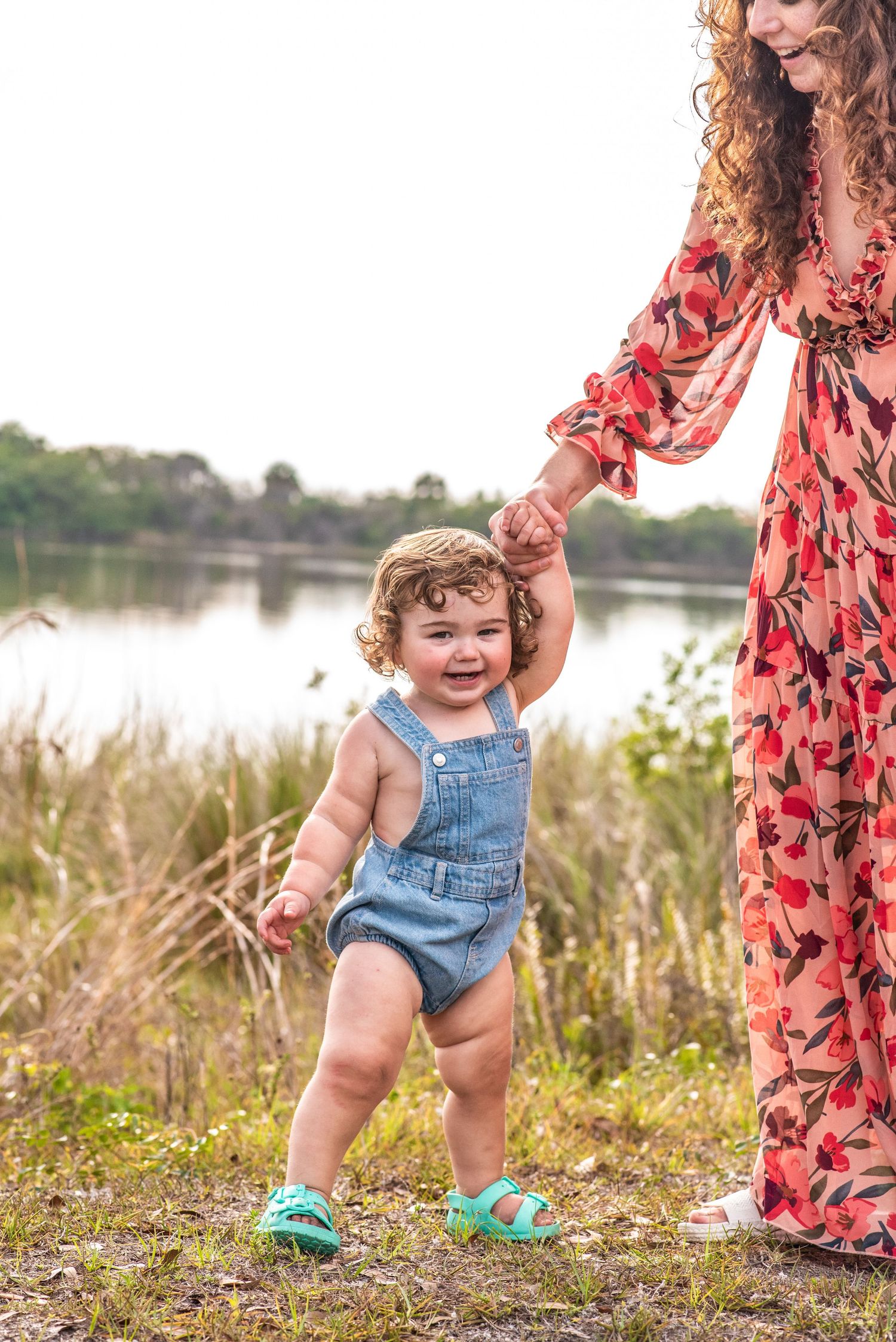 A smiling toddler in denim overalls holds a woman's hand while learning to walk near a scenic lake.