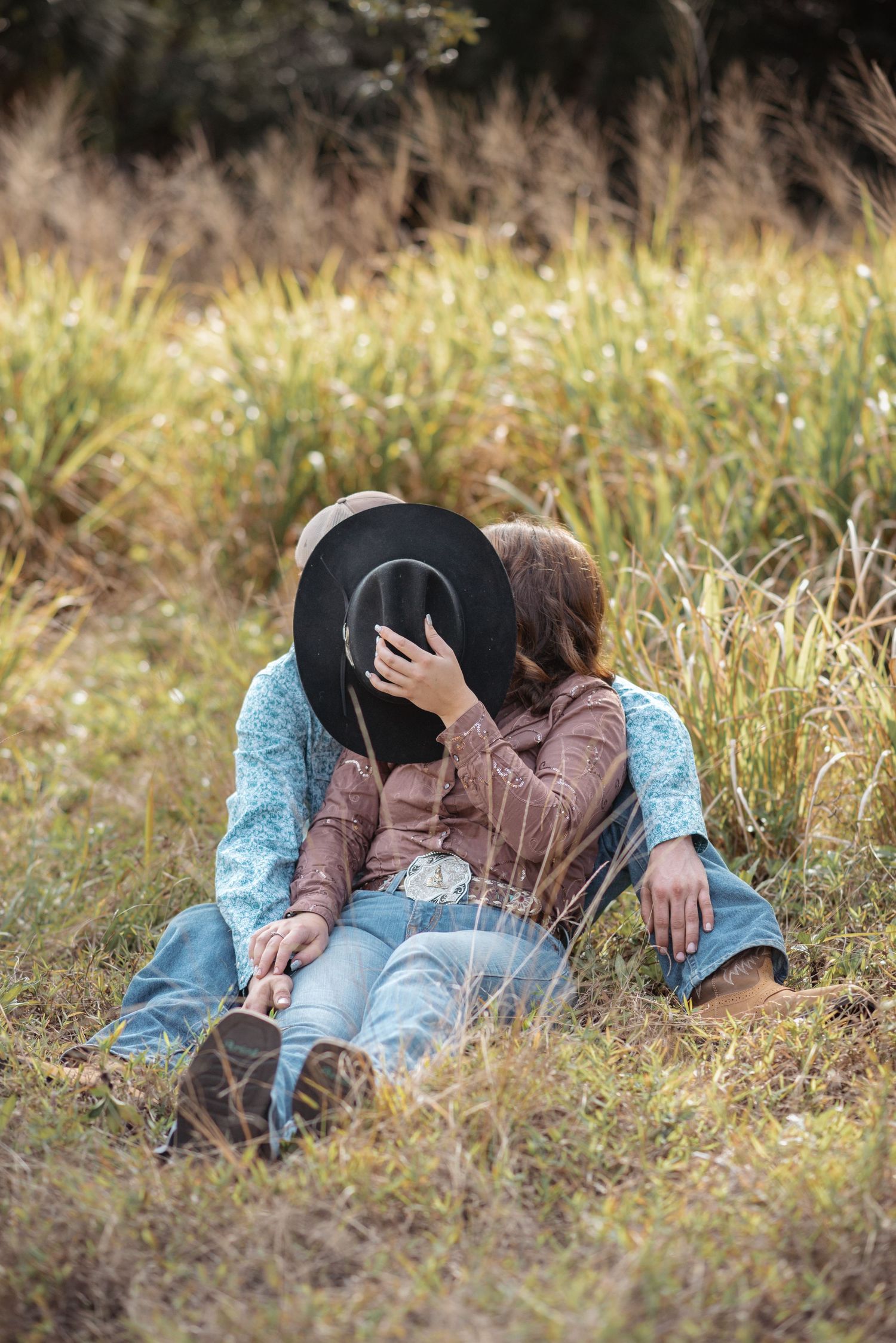 Couple sitting in a grassy field, sharing a kiss hidden behind a black cowboy hat, wearing jeans and western shirts.