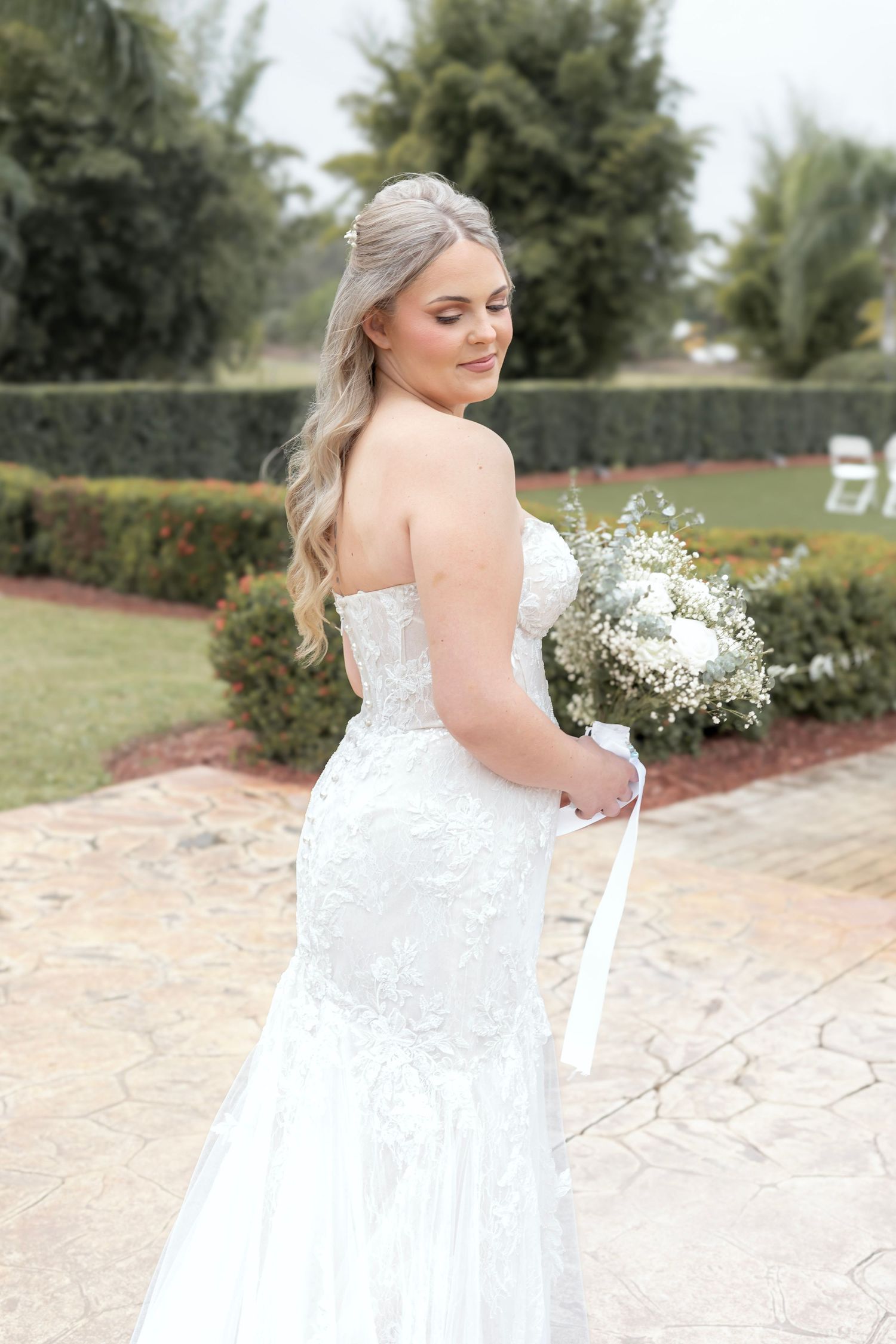 Bride in strapless lace mermaid gown holding white bouquet, posing outdoors on stone pathway.