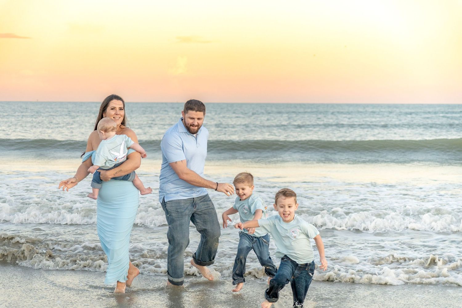 Family of four playing in ocean waves at sunset on a sandy beach, parents holding children's hands.