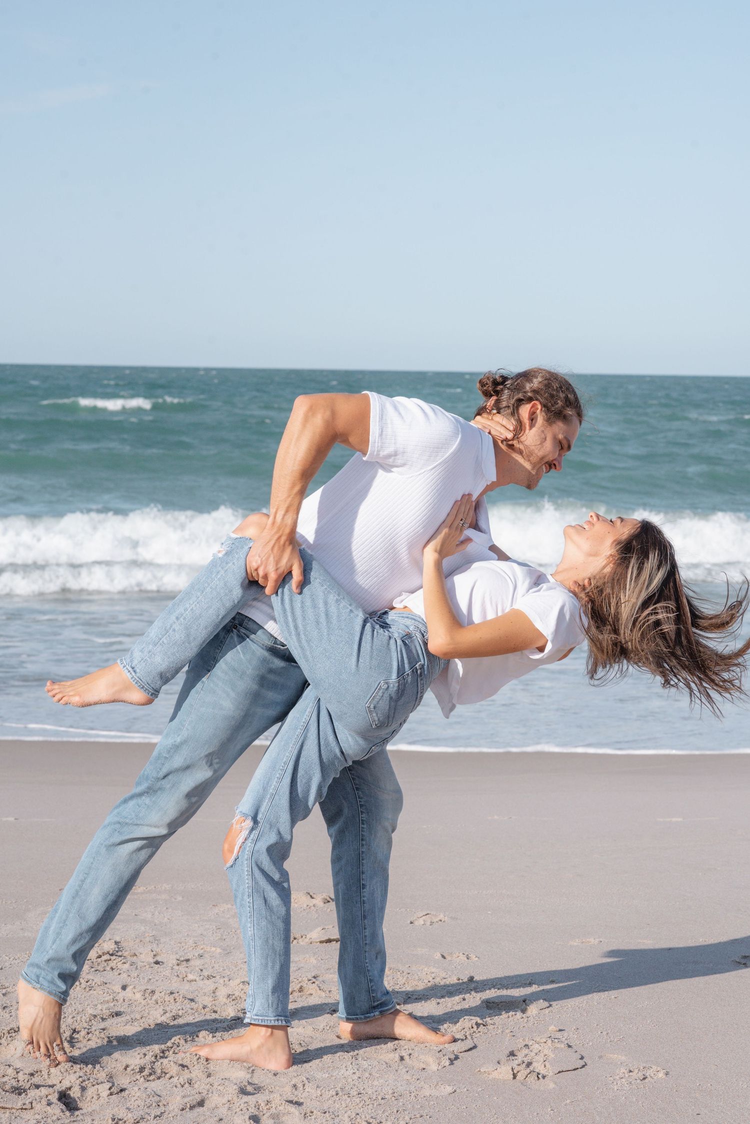 Couple dancing romantically on a sunny beach, man dipping woman back near ocean waves, both wearing white shirts and jeans.