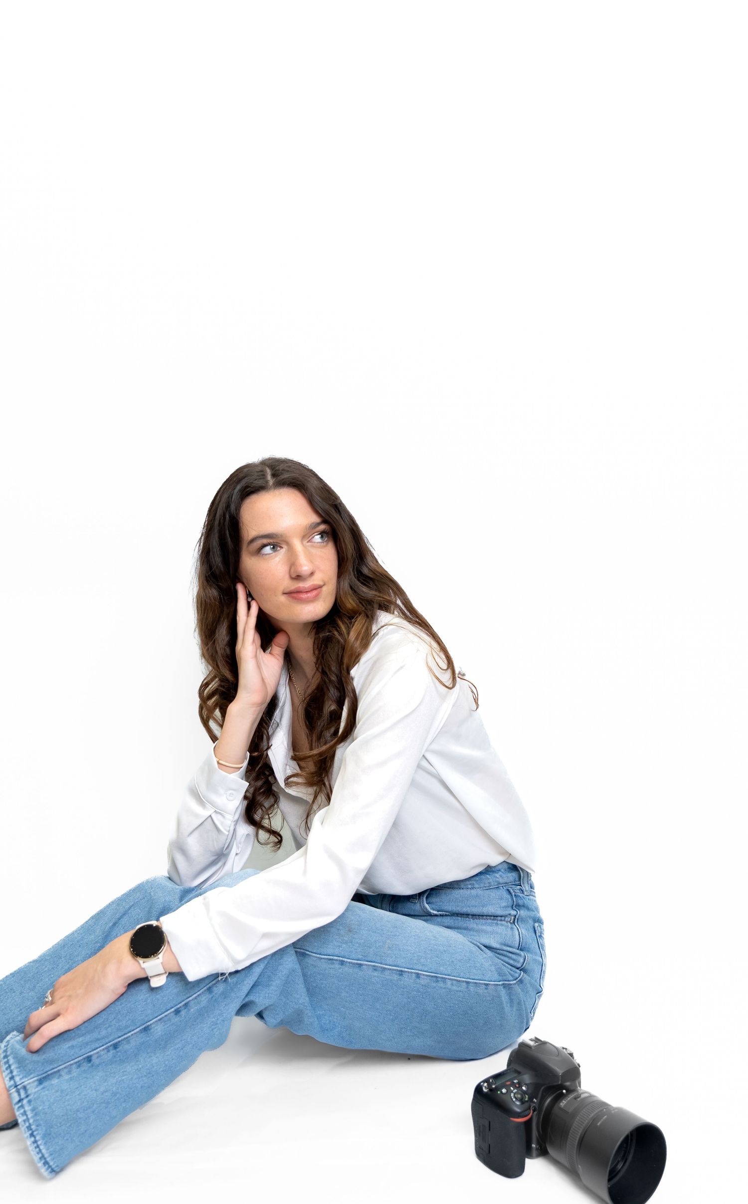 Young female photographer sitting on floor in white blouse and jeans, looking up thoughtfully with camera beside her.