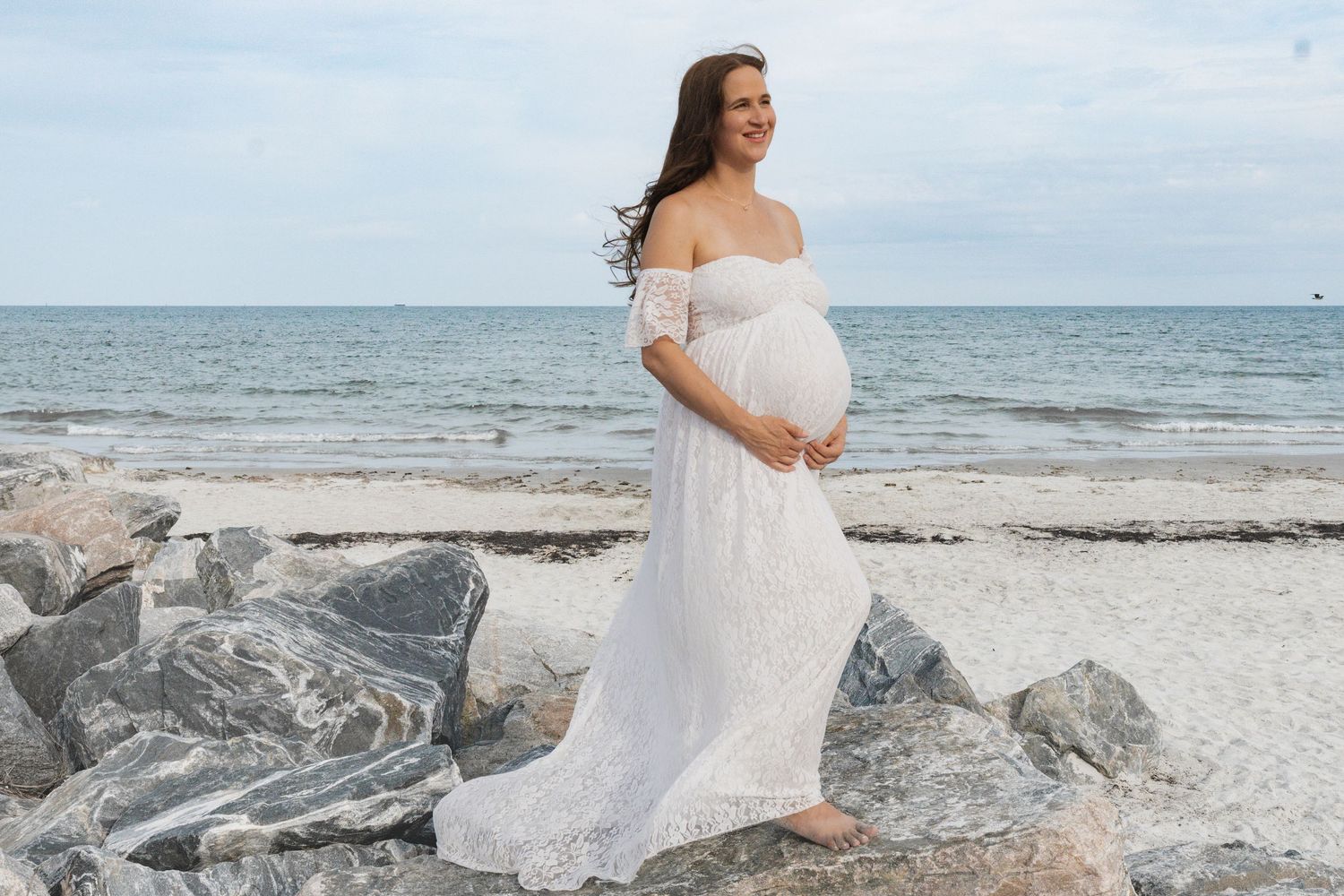 Pregnant woman in white lace off-shoulder gown posing on rocky beach by the ocean.