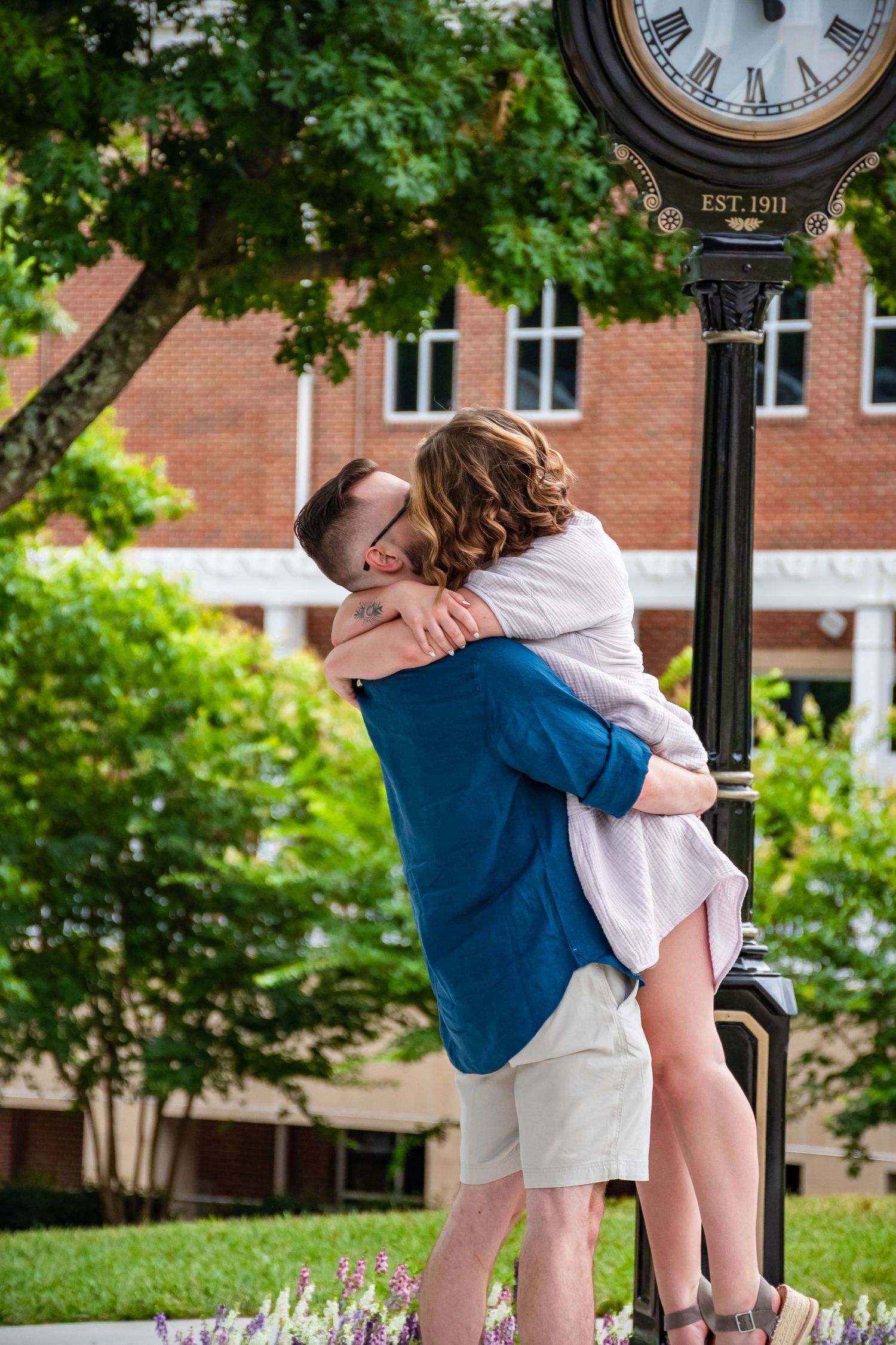 Couple embracing romantically near an EST. 1911 clock tower on a college campus surrounded by lush greenery.
