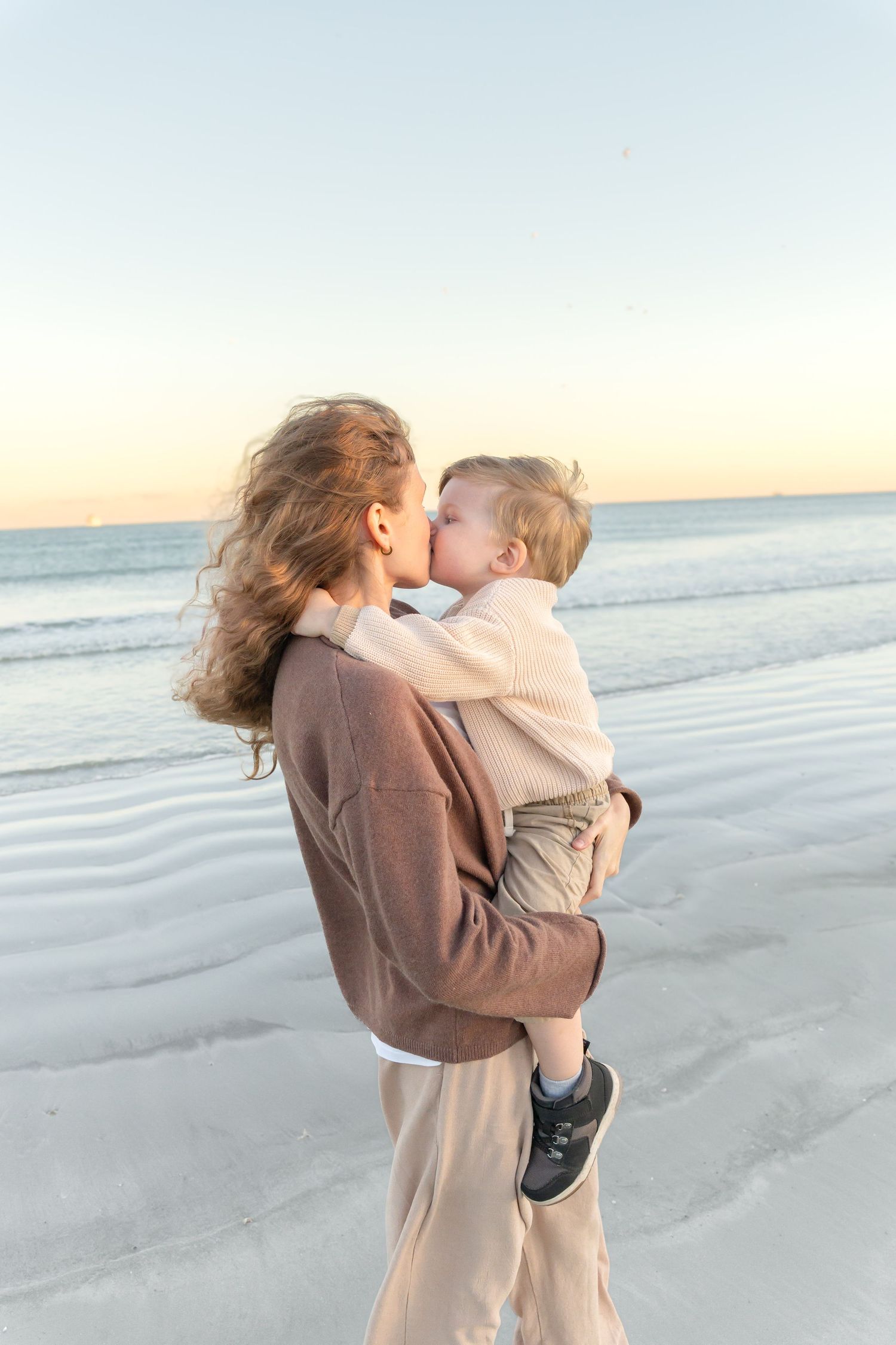 A mother kisses her toddler while holding them on a serene beach at sunset.