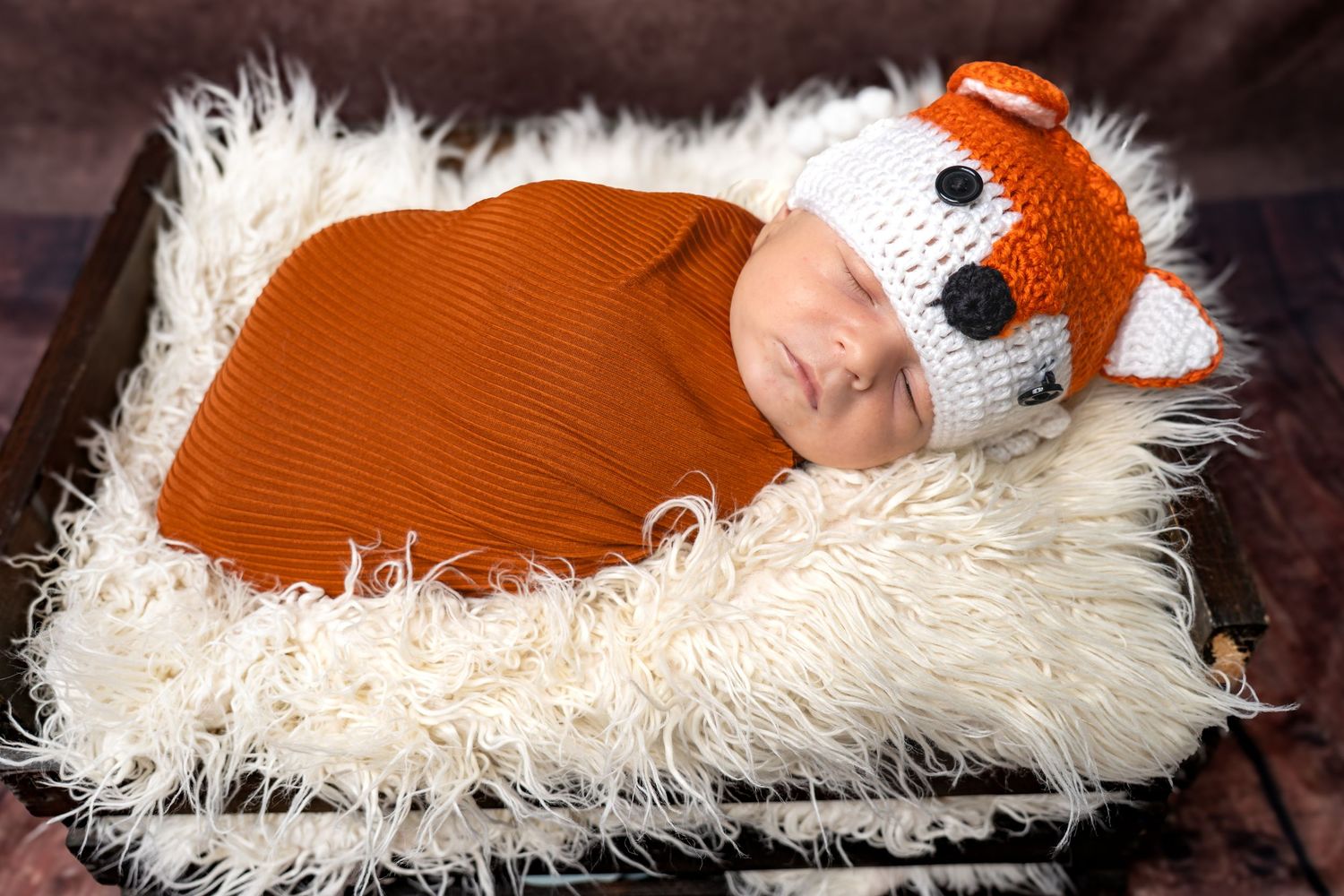 Newborn baby sleeping in a wooden crate wrapped in orange swaddle wearing a crocheted fox hat on fluffy white rug.