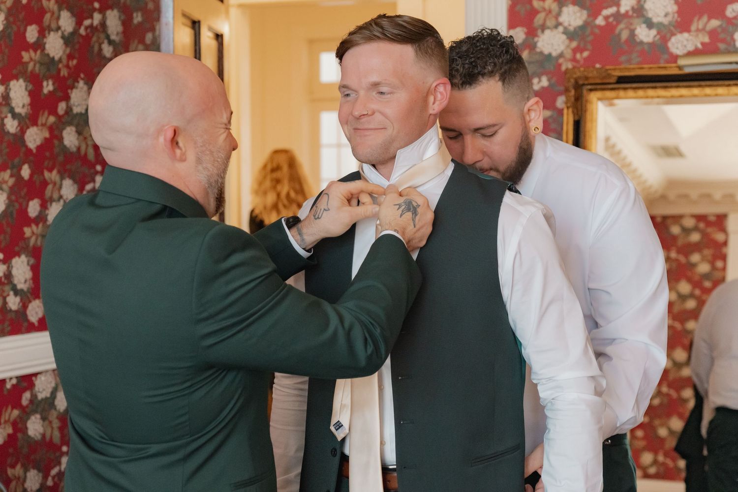 Two groomsmen in dark green suits help adjust the groom's white tie before the wedding ceremony.
