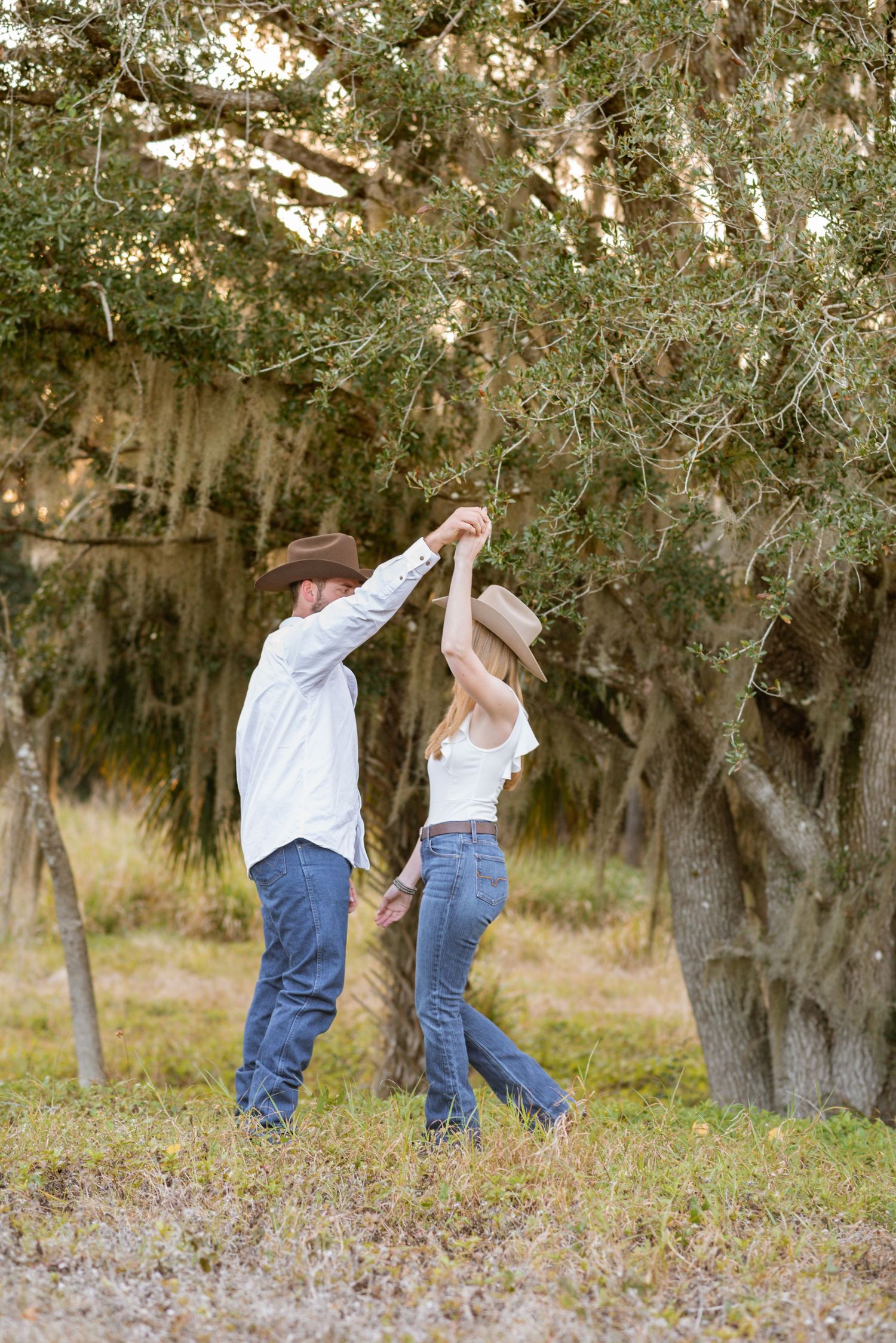 Couple in cowboy hats dancing under a large oak tree in a rustic outdoor setting, wearing jeans and white tops.